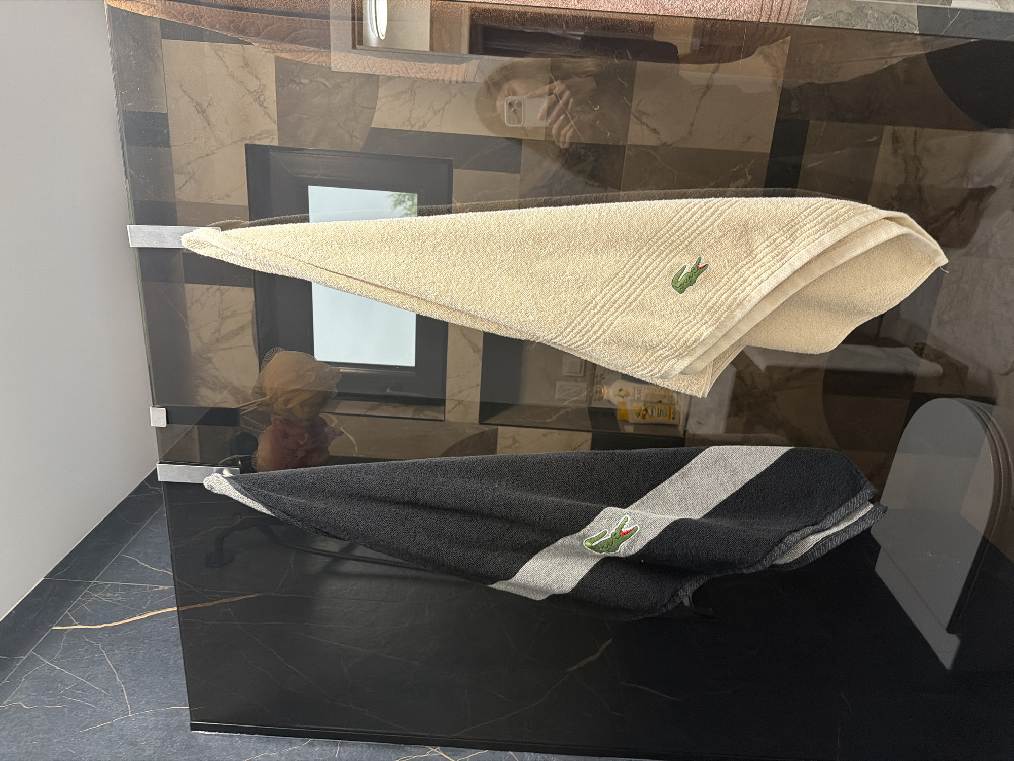 A photograph depicts two towels on a dark, reflective surface, potentially a countertop or shelf. The towels are branded with the Lacoste logo, one in a light beige color and the other in a black and gray stripe. The beige towel is hanging from a metal rod, while the black and gray towel rests on the surface below.
The background features a brown and gray geometric pattern on the wall. There is also a small, rectangular window or mirror in the wall's background. A faint reflection of a person holding a phone is visible in the upper portion of the reflective surface.
The lower left corner reveals a black tiled floor with golden veining.
The location in the city of Resilovo, Bulgaria, cannot be determined from the visual information alone.