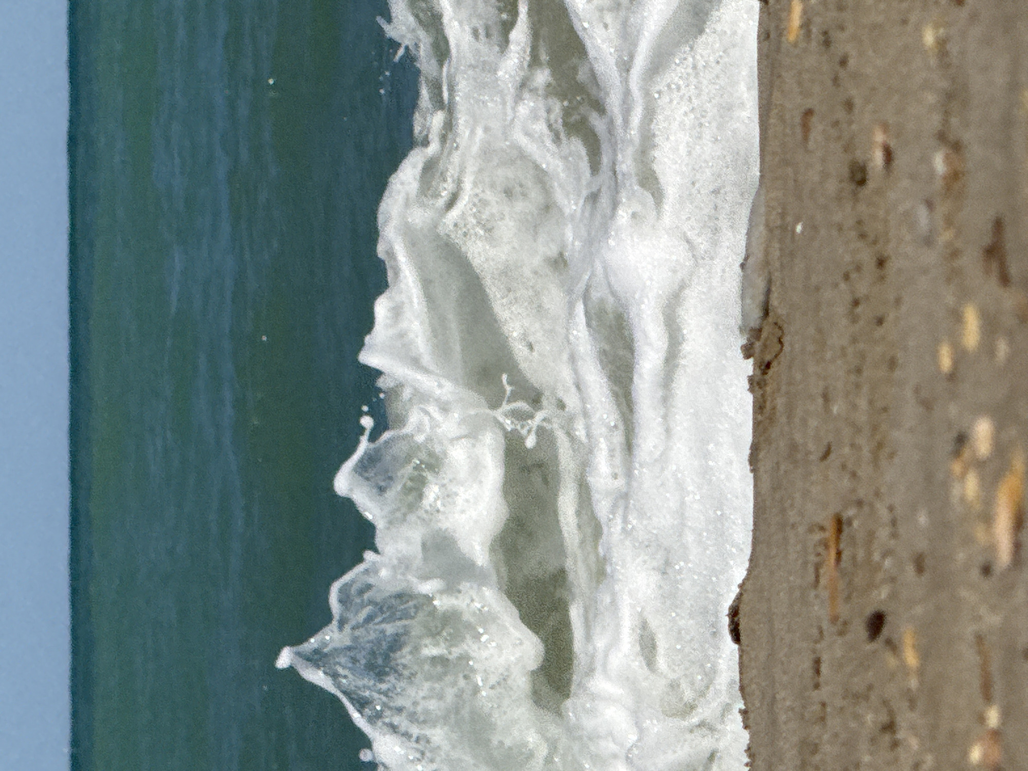 A close-up, eye-level shot captures a wave crashing on a sandy beach. The water is a deep teal, and the sky above is a pale blue. The wave is white and foamy, with a few droplets of water splashing upwards. The sand in the foreground is light brown and appears to be composed of small pebbles and shells. The image does not depict any people or structures, and its location in San Benedetto del Tronto, Italy, is not discernible from the visual elements alone.