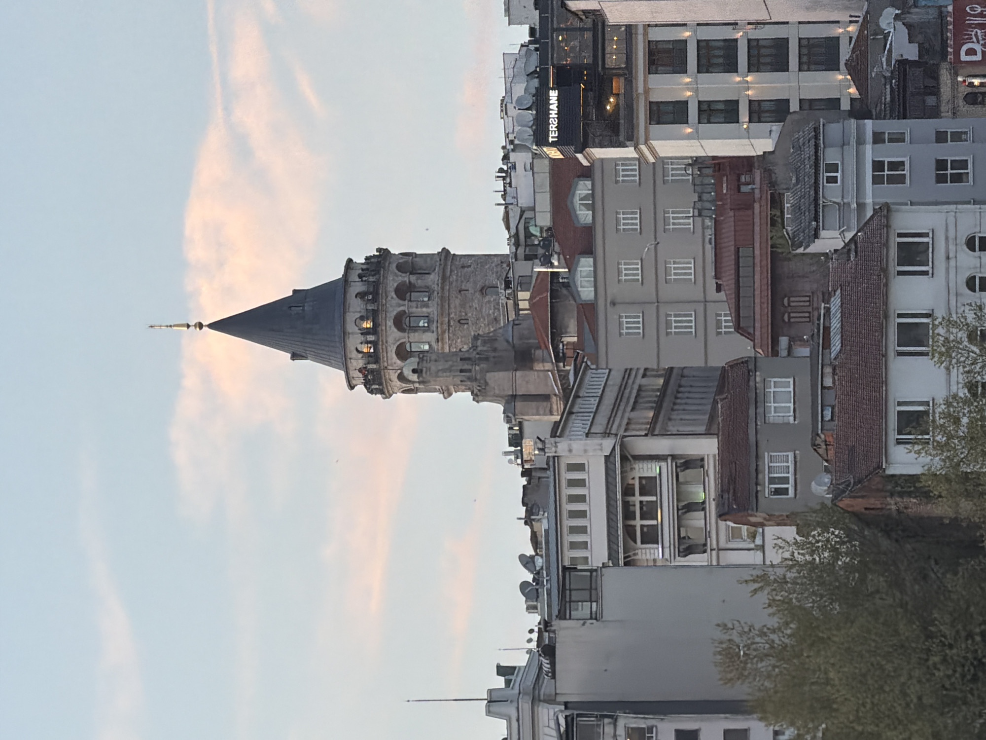 The image displays a prominent view of the Galata Tower in Istanbul, Turkey, set against an evening sky. The historic stone tower, located on the left, is cylindrical with a distinctive dark, conical spire. Its upper levels feature arched windows and balconies, with warm interior lights illuminated, and several small figures visible, likely tourists, on an observation platform.
To the right of the Galata Tower, a dense array of urban buildings fills the frame. These structures vary in height and architectural style, ranging from modern multi-story buildings with numerous windows to older constructions with tiled rooftops. Many rooftops are equipped with satellite dishes. One building prominently features an illuminated sign displaying the word "TERSHANE" (likely 'TERSANE').
The sky is a soft, clear blue, adorned with delicate streaks of light pink and orange clouds, indicative of either a sunrise or sunset. Given the artificial lights glowing from the tower and the "TERSHANE" sign, it most likely depicts the onset of twilight or early evening. The overall scene captures a vibrant urban environment, with the Galata Tower standing as a key landmark in the Beyoğlu district of Istanbul. No specific active event is depicted beyond the general ambiance of the city at dusk.