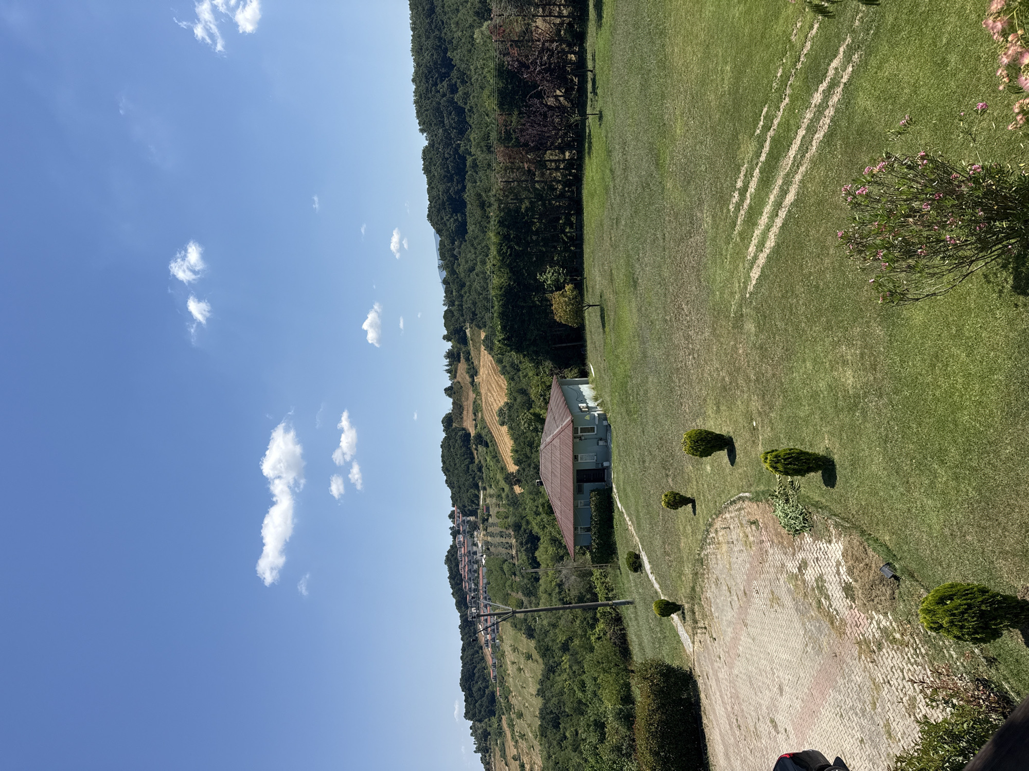 The image depicts a scenic view of a rural landscape under a clear blue sky with scattered white clouds. In the foreground, a well-maintained grassy area leads to a small, light blue house with a reddish-brown roof. A pathway made of interlocking pavers is visible to the left, with a dark-colored vehicle parked partially in frame. Several small, conical evergreen bushes are strategically placed around the property.
The middle ground features a dense line of tall trees, with a variety of foliage colors suggesting different species. Beyond the trees, the landscape rises into rolling hills covered in a mix of green trees and cultivated fields, some appearing to be dry or harvested. In the far distance, a cluster of white houses with red roofs are nestled on a hillside, overlooking the valley. A utility pole stands prominently in the mid-ground, near the house. The overall scene conveys a sense of tranquility and natural beauty, characteristic of the Termal region in Turkey.