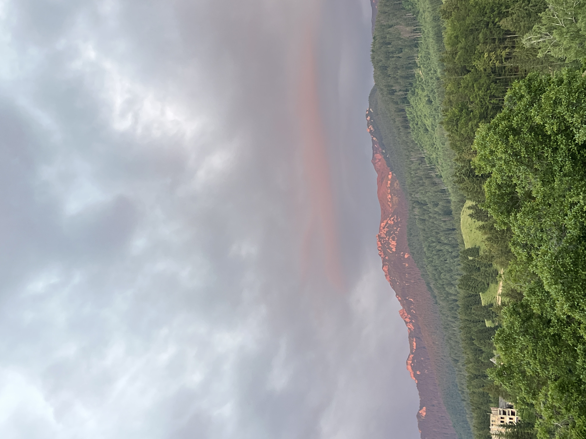This is an eye-level, full shot of a mountainous landscape under a cloudy sky. The top two-thirds of the image is dominated by a grey, overcast sky with hints of pink and orange light reflecting off the clouds near the horizon. Below the sky, a rugged mountain range stretches across the frame. The upper slopes of the mountains are rocky with sharp peaks, catching a warm, pinkish light from the setting or rising sun. These peaks are in stark contrast to the dark green, densely forested lower slopes that descend into a valley. In the foreground, lush green trees, some with lighter green leaves, create a dense canopy. To the far left, a multi-story, unfinished building with visible concrete floors and exposed rebar stands amidst the trees. The overall scene depicts a natural mountain environment with signs of human development.
