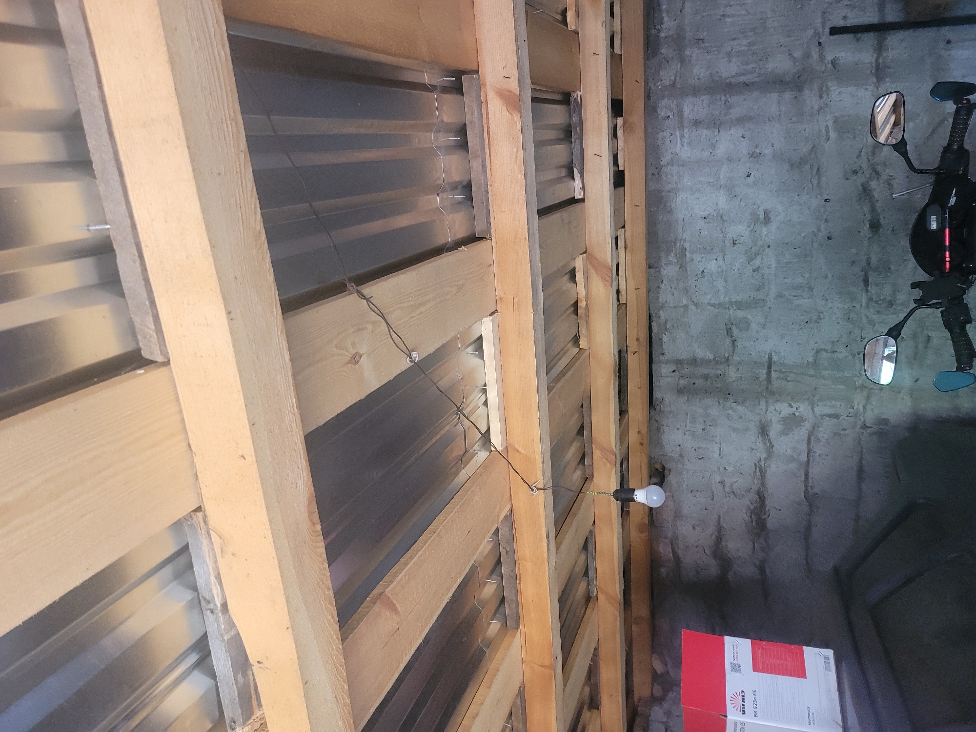 The image depicts the interior of a garage or workshop in Vovkivka, Ukraine. The perspective is looking upwards at the ceiling construction. The ceiling is made of corrugated metal sheeting supported by wooden beams and planks. Some of these planks are unfinished wood, showing their natural grain. A single lightbulb hangs from a wire suspended from the ceiling.
In the lower portion of the image, the concrete wall of the garage is visible. To the left, a portion of a cardboard box with red and white branding is present. In the foreground, the handlebars and front section of a black scooter are visible, suggesting the space is used for vehicle storage or maintenance. The overall impression is one of a functional, perhaps unfinished, workspace.