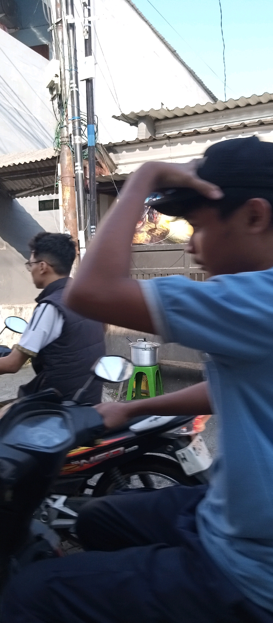 In Wadungasri, Indonesia, two young men are on a motorcycle. The man in the foreground, wearing a blue t-shirt and a black cap, is facing away from the camera, reaching up to adjust his cap. The man behind him, wearing glasses and a black vest over a striped shirt, is also on the motorcycle. The motorcycle is in motion, indicated by the blur. In the background, there is a white building with a corrugated metal roof, electrical poles with tangled wires, and a green plastic stool with a metal pot on top. A poster of a motocross rider is visible on the wall behind the stool. The sky is clear and blue.