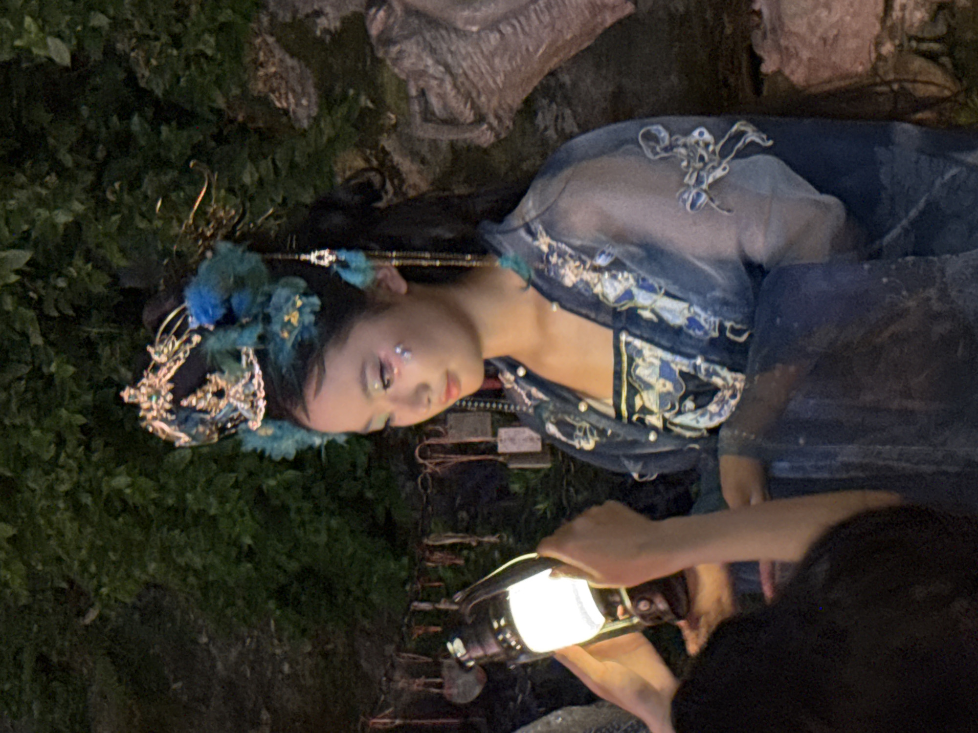 A young East Asian female, adorned in a traditional Chinese costume with elaborate blue and gold headdress and matching garments, holds a lit lantern. Her makeup includes glittery accents on her cheeks and eyes. She stands in what appears to be an outdoor, possibly ancient, setting, with lush green foliage forming the background and rocks visible to the right. The scene suggests a cultural event or performance, possibly in the Yuzhong District of China, known for its historical sites. A blurry dark shape in the foreground may be another person or object, and another hand is visible holding the lantern. Various hanging objects, possibly prayer charms or tokens, are strung up behind the subject.