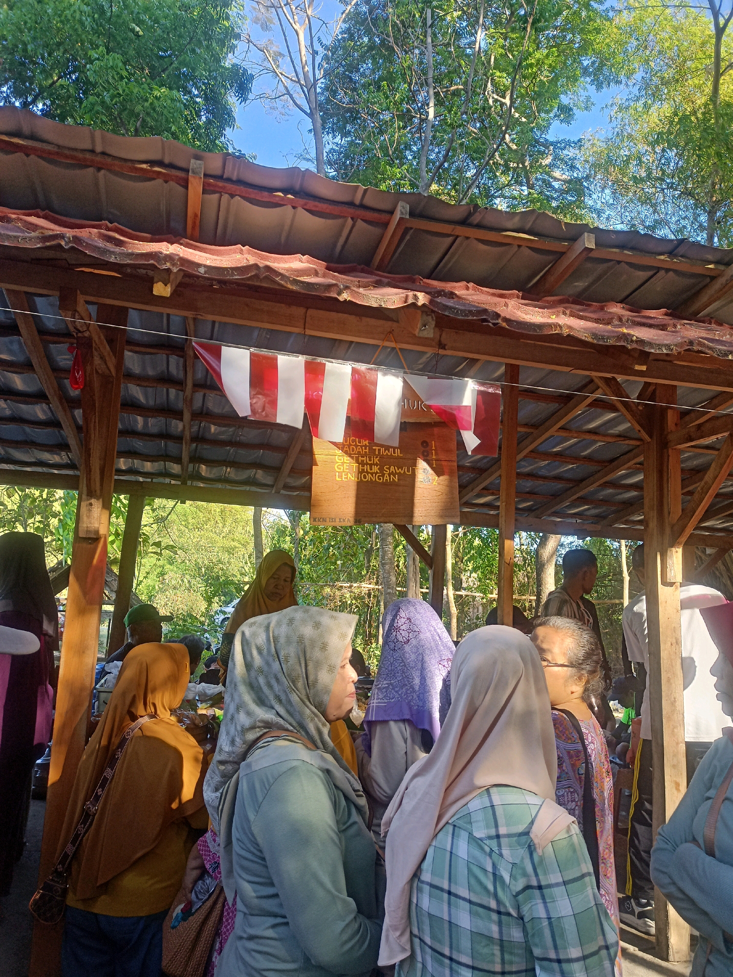 The image captures an outdoor scene under a wooden, open-sided shelter with a corrugated metal roof, likely in the city of Karungan, Indonesia. A group of people, predominantly women wearing hijabs, are gathered. The shelter's roof is supported by wooden beams and posts. Red and white Indonesian flags are displayed horizontally under the roof. A wooden sign with text in Indonesian is also visible. The background shows lush green trees and a bright blue sky, indicating an outdoor, possibly rural or semi-rural, setting. The people appear to be engaged in some form of gathering or transaction, with some looking towards the interior of the shelter. The lighting suggests it is daytime, with sunlight filtering through the trees.