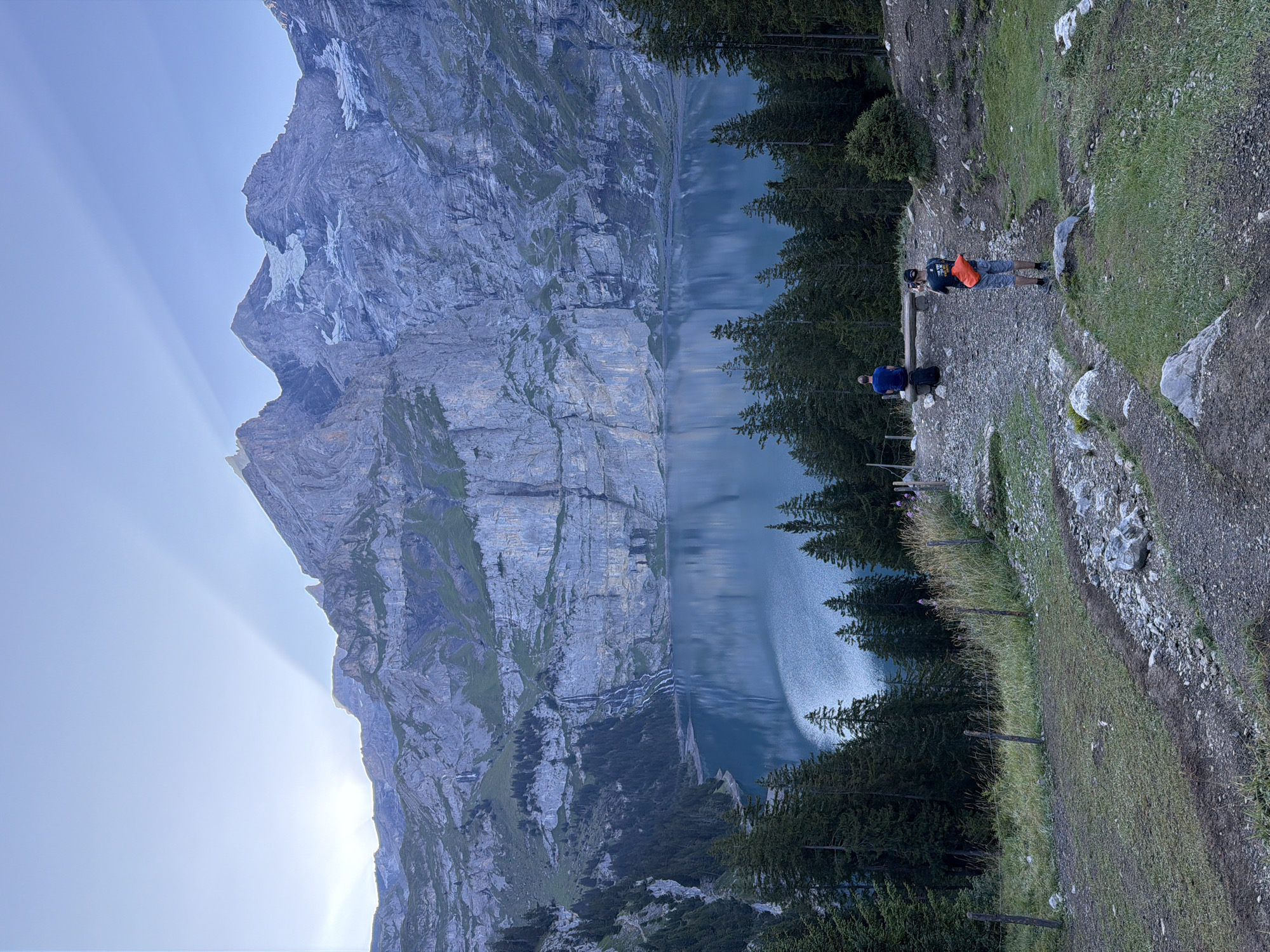 An outdoor, vertical, full shot captures a scenic view of the Oeschinensee lake and surrounding mountains in Kandersteg, Switzerland, during twilight. A crystal-clear, blue lake reflects the rugged, grey cliffs and green trees that line its shore. Waterfalls cascade down the sheer rock faces, adding to the dramatic landscape. In the foreground, a gravel path winds through patches of grass, with two men positioned on the path. One man, wearing a blue shirt, sits on a wooden bench facing the lake, with a black backpack beside him. The other man, wearing a grey t-shirt and shorts, stands facing away from the camera, holding a phone to take a picture of the scenery, with an orange fanny pack around his waist. A wooden fence with wire stretches across the left side of the image, demarcating a grassy area. The sky above is a pale blue, with ethereal light rays fanning out from behind the sun, partially obscured by the jagged mountain peaks.