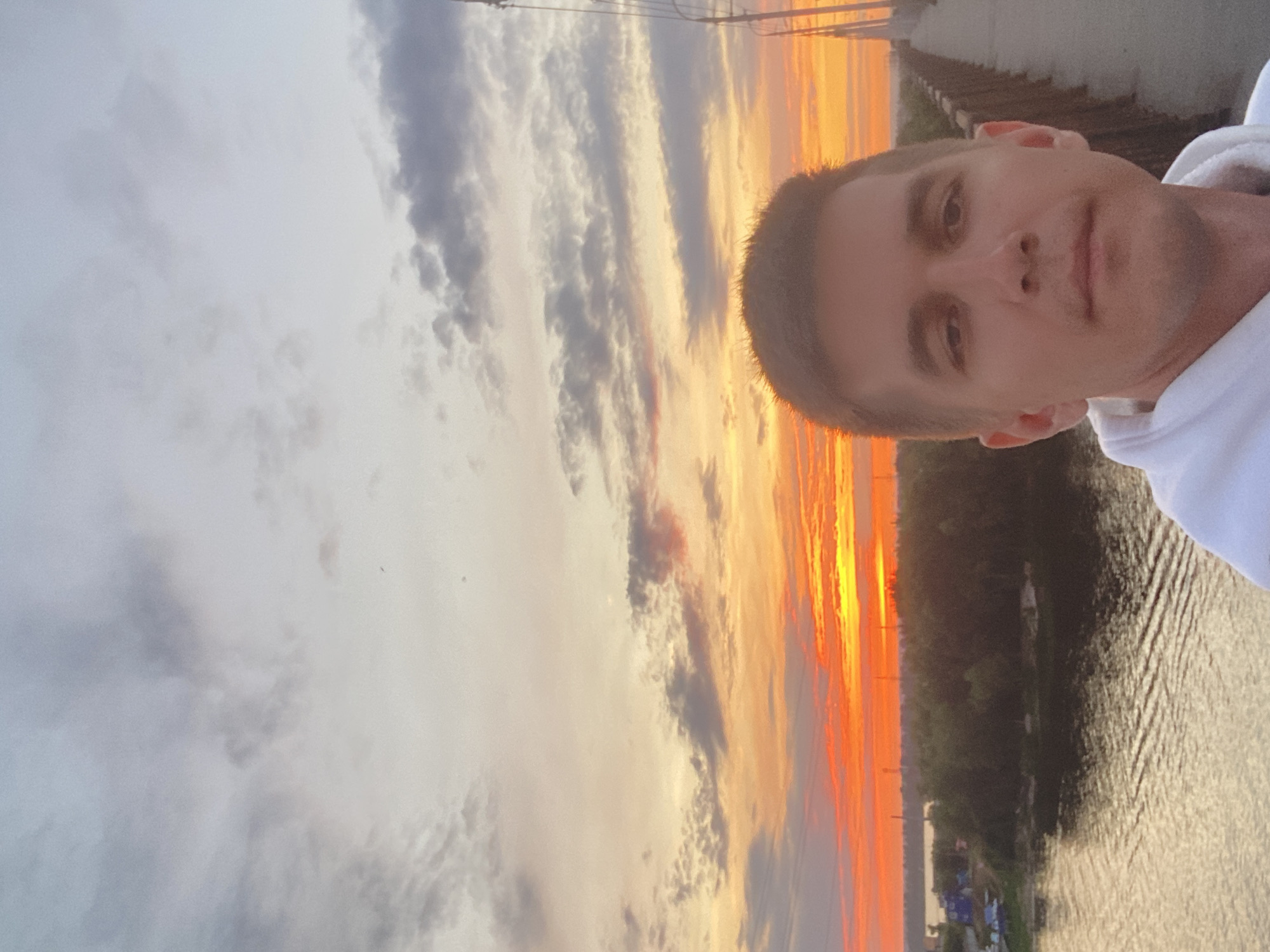 A selfie is taken by a man in the foreground, his head and shoulders filling the lower right of the frame. He is wearing a white hooded sweatshirt. The man is standing on a bridge, with a railing visible to his right. The bridge spans a body of water, likely a river, which occupies the lower left of the image. The water's surface is rippled. Beyond the river, a treeline is visible, with some structures and industrial elements peeking through. The sky is dominated by a vibrant sunset, with shades of orange, yellow, and red clouds creating a dramatic backdrop. The location is identified as Novokuybyshevsk, Russia.