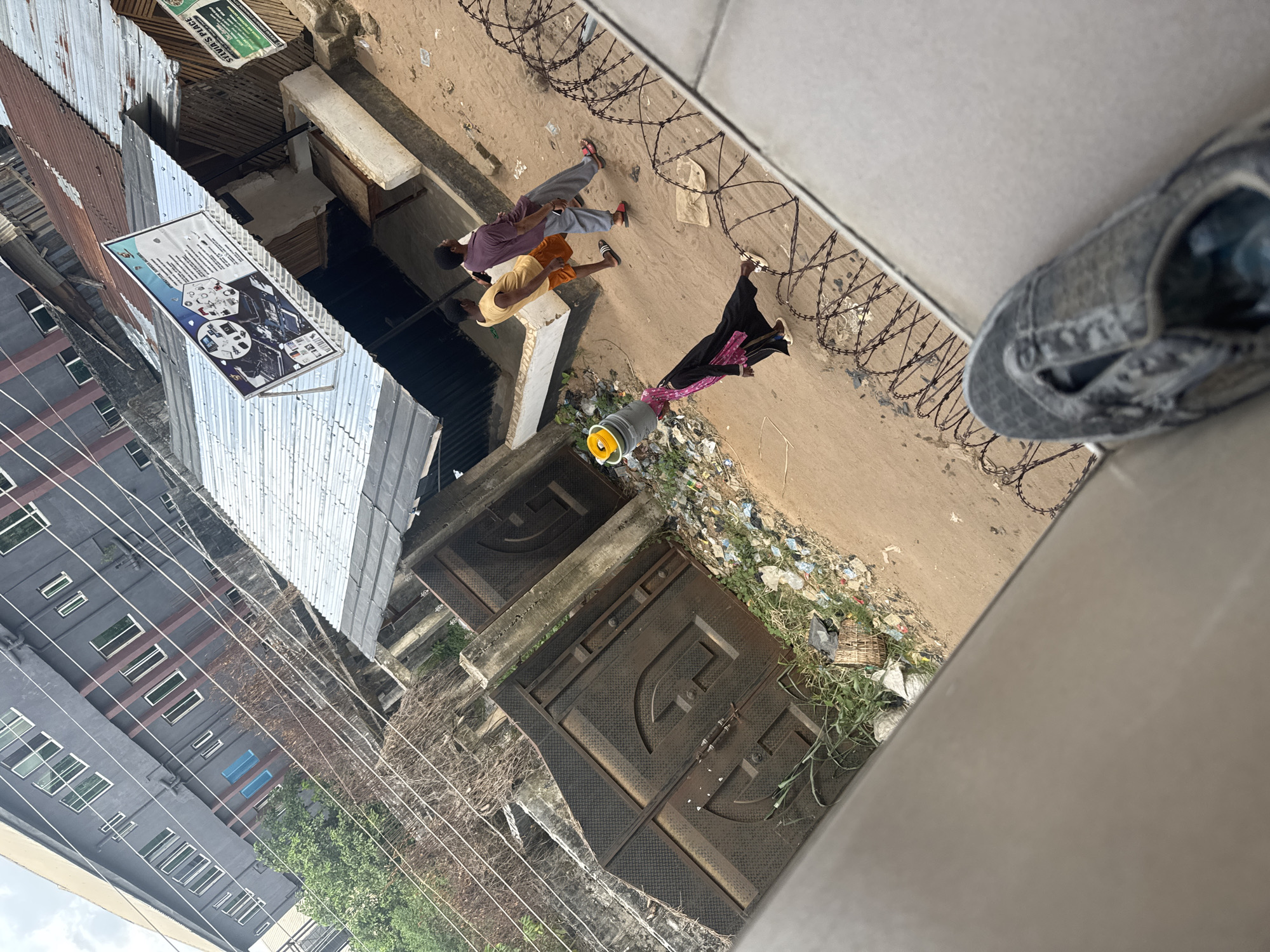 An eye-level shot, taken from above, of a street scene in Ihiagwa, Nigeria. Three pedestrians are visible: two men and a woman in a dark, full-body dress. The men are wearing casual clothes. Barbed wire lines the edge of the street. Rubbish, litter, and sparse vegetation cover the area adjacent to the street. There are two metal gates on the side of the street. One gray building is visible. The sky is overcast. A sign is visible. A shoe can be seen in the foreground.