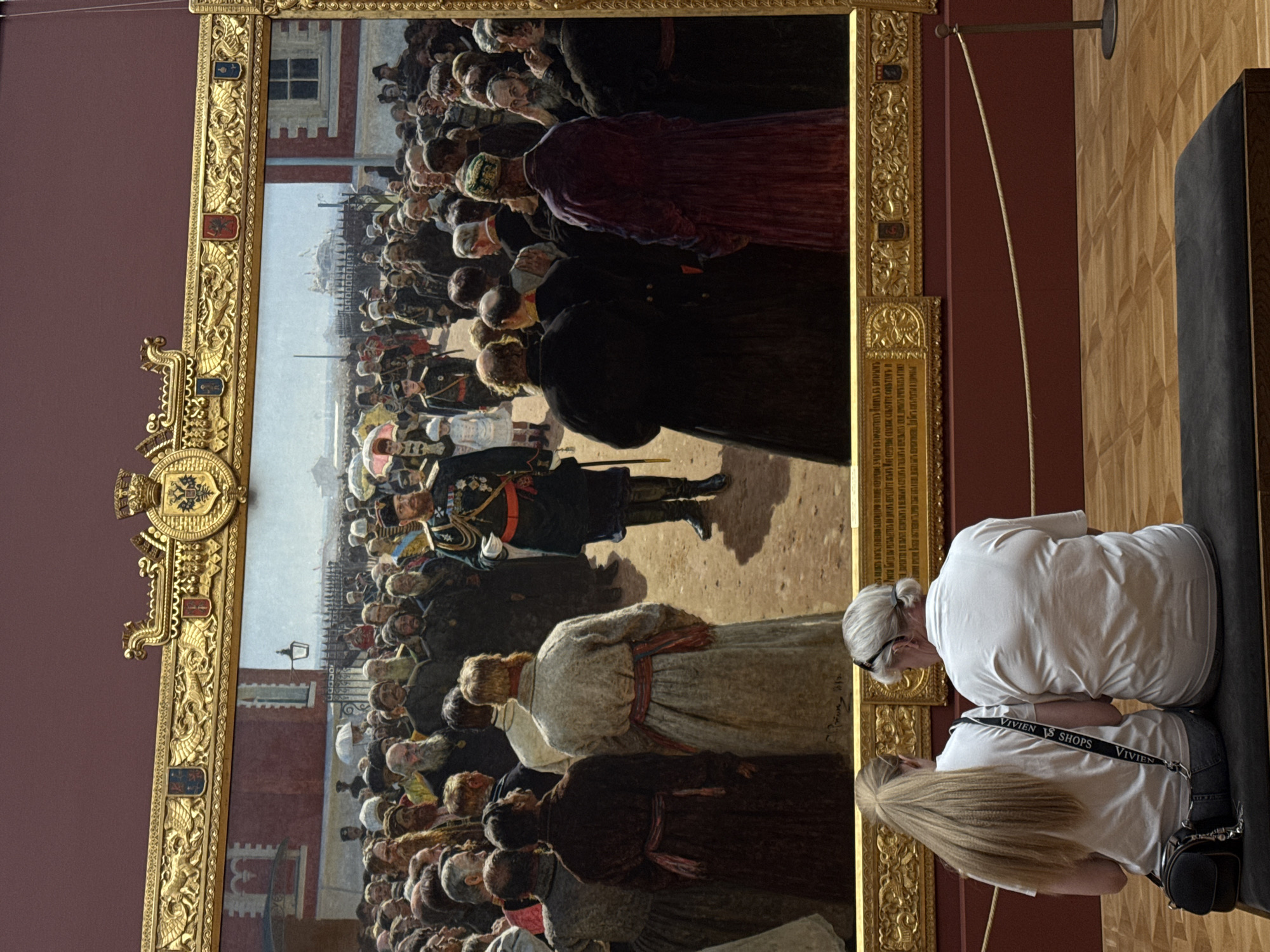 A photo of a large historical painting in a gold frame depicting a crowd of people in what appears to be a procession or ceremony. The painting seems to depict a city, possibly Moscow, with buildings and structures in the background. In the foreground, in front of the painting, there are two people sitting on a bench. The background is a maroon color.