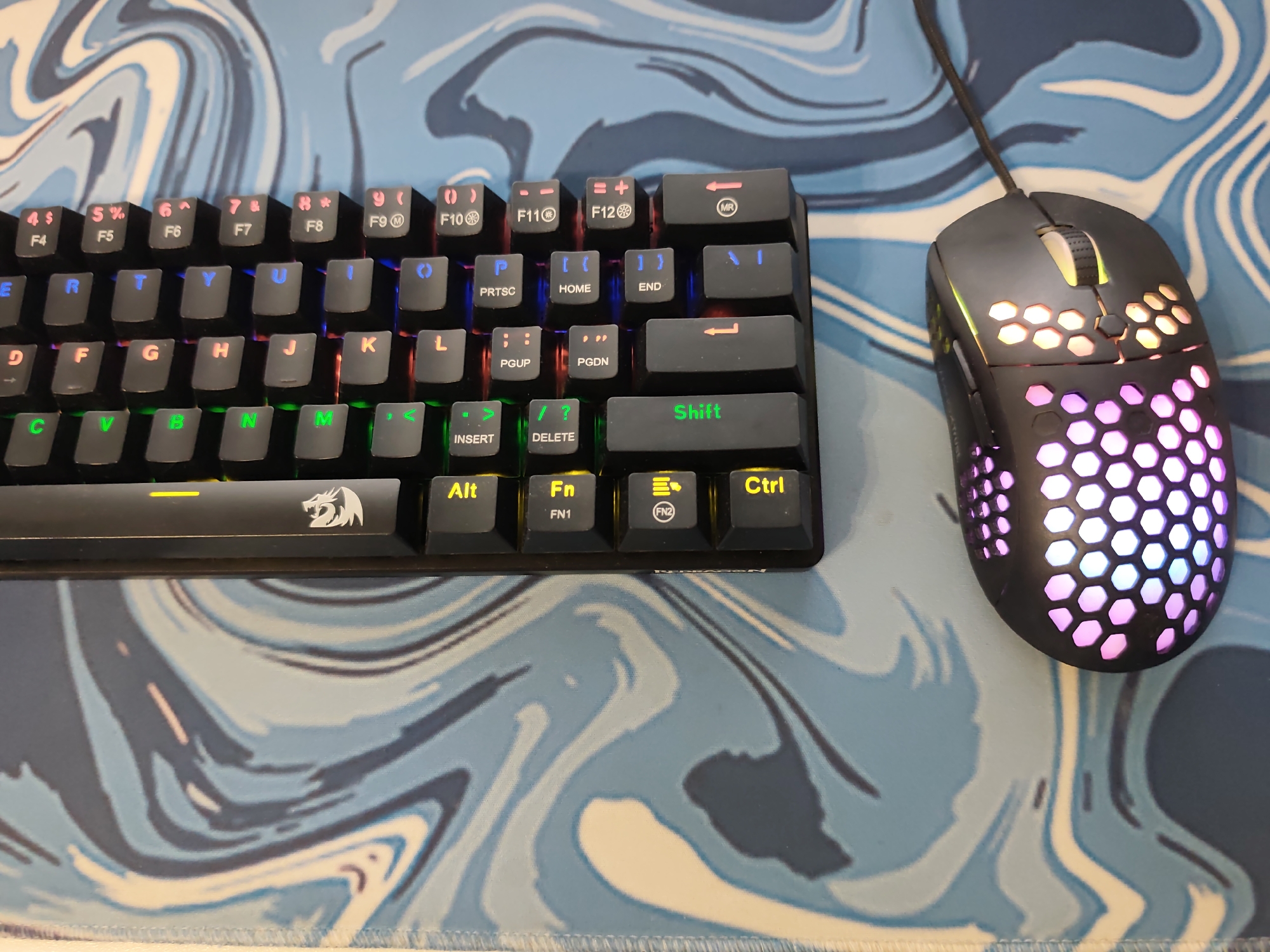 The image presents an overhead view of a computer keyboard and a gaming mouse situated on a patterned desk mat. The keyboard is a black mechanical model with RGB backlighting, illuminating its keycaps in multiple colors: pink on the F-row number keys (F4-F10), blue on the QWERTYUI keys, orange/red on the ASDFGHJK keys, and green on the ZXCVBNM keys. The spacebar features a visible Redragon logo. The mouse is also black, characterized by a honeycomb shell design that allows internal RGB lighting to glow through. This lighting transitions from a warm yellow/orange at the front near the scroll wheel to purple and blue hues towards the rear. A black braided cable extends from the mouse's front. Both peripherals rest on a fabric desk mat adorned with an abstract, swirling pattern in varying shades of blue and white. There is no visual information within the image to determine a specific geographical location, such as Pazardzhik, Bulgaria.