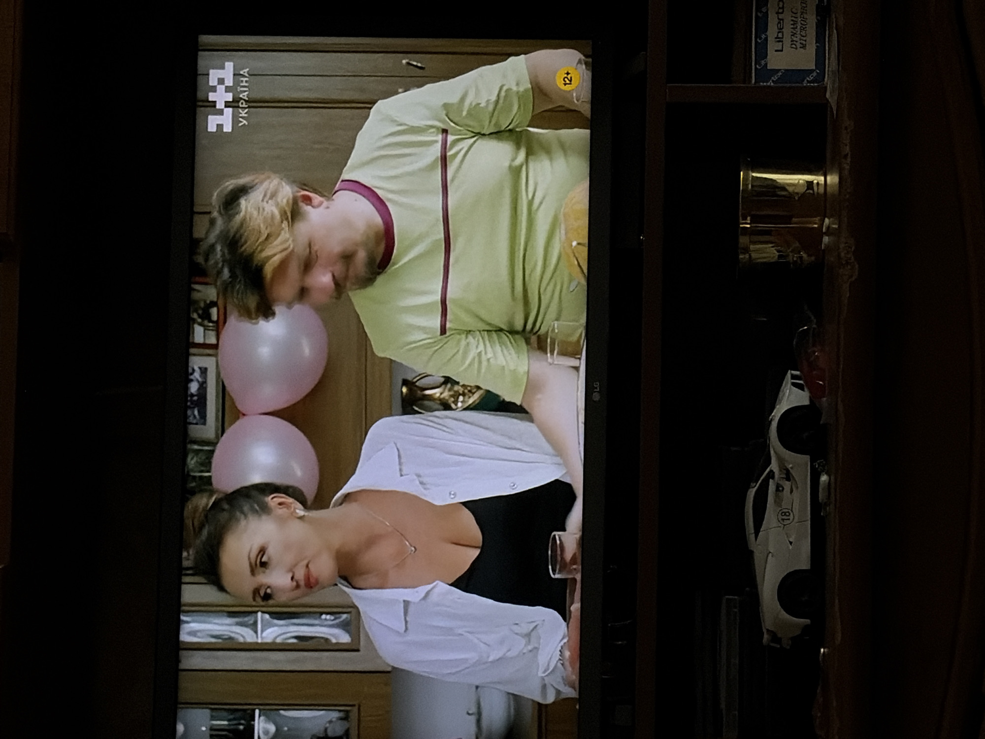 On a television screen, a man and a woman are seated at a table in what appears to be a home setting. The woman, on the left, is wearing a white open shirt over a black top and a necklace. The man, on the right, is wearing a light green t-shirt with a maroon trim. Behind them, there are two pink balloons and a logo indicating "1+1 UKRAINA". A "12+" rating is visible in a yellow circle on the right side of the screen. The lower part of the image shows the top of a dark entertainment unit with a white toy car and a golden cylindrical object on a shelf. The scene is framed by the edges of the television screen.