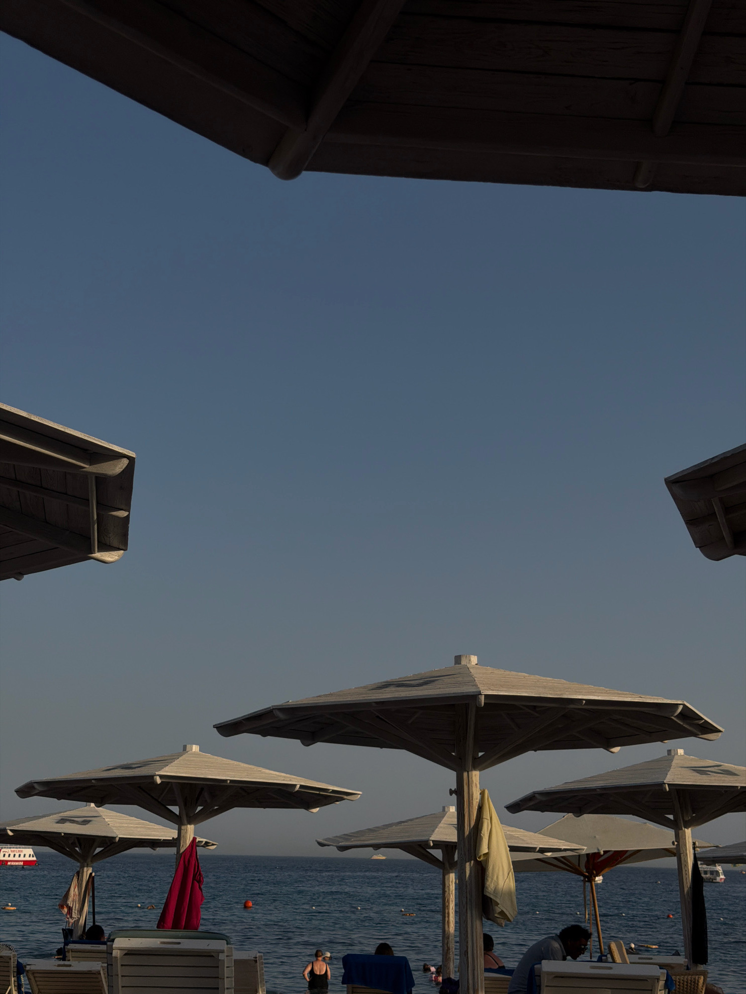 This is a photograph of a beach scene in Sharm El Sheikh, Egypt, likely taken in the late afternoon. The image is framed by the wooden structures of beach umbrellas in the foreground. The sky is a clear, pale blue. In the middle ground, several large, hexagonal beach umbrellas with light-colored fabric tops are set up on a sandy beach. Two of these umbrellas have towels or clothing draped over their poles, one a bright red and the other a pale yellow. In the background, the calm, blue sea stretches to the horizon. A white and red tourist boat is visible on the water to the left, and several smaller buoys are scattered on the surface. Some lounge chairs are visible on the beach, and a few people are present, some sitting and some standing near the water's edge. A woman in a black swimsuit is standing by the water on the right. The overall impression is one of a tranquil beach day.