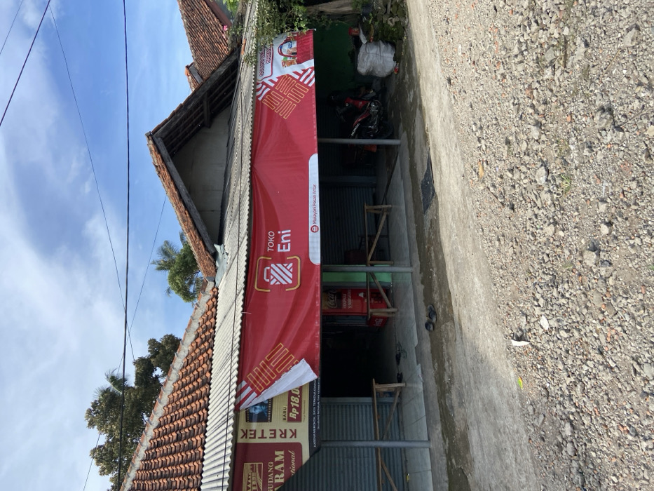 The image shows a small storefront in Sukatani, Indonesia, located at the coordinates -6.643 latitude and 107.414 longitude. The storefront is a simple, one-story building with a corrugated metal roof and a red banner above the entrance. The banner advertises a shop called "Toko Eni," with a slogan that reads "Melayani Pesan Antar." The storefront has two open doorways and a single window. A red Coca-Cola cooler is visible on the left side of the storefront. A motorcycle is parked on the right side of the building. There are two wooden benches in front of the storefront. The ground in front of the building is covered in gravel. There is some vegetation visible in the background. The sky is blue with some white clouds. There are no people visible in the image.