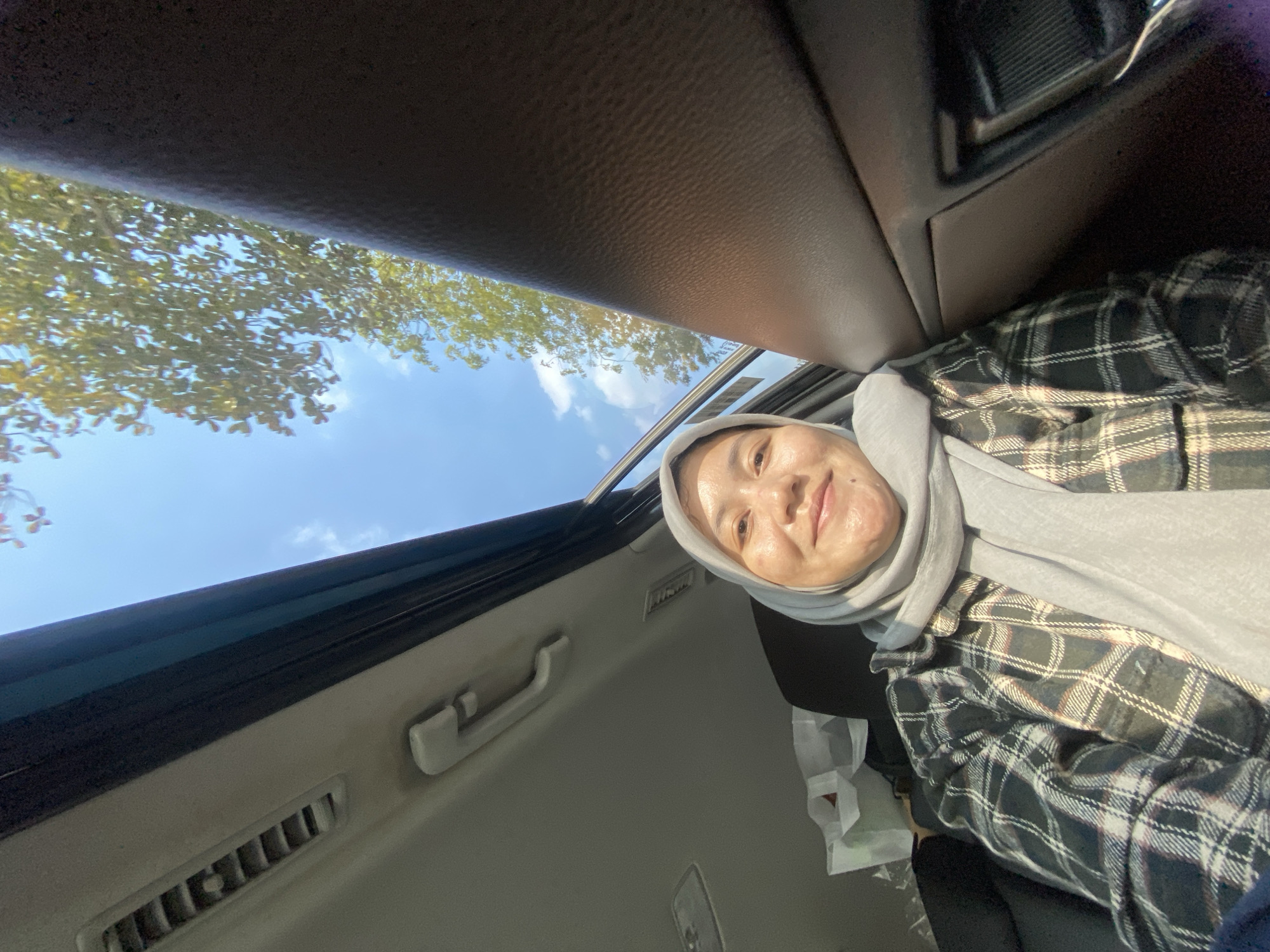 A woman wearing a light gray hijab and a black and white plaid shirt is captured in a selfie taken from inside a vehicle, looking upwards towards the open sunroof. The sunroof reveals a bright blue sky with scattered white clouds and the leafy branches of a tree. The interior of the car is visible, including the car's ceiling with an air vent and a handle, and the textured brown door panel. The woman is smiling and appears to be enjoying the sunny weather. The setting appears to be a street in Banjarmasin, Indonesia, given the sunny climate and lush vegetation typically seen in the region.