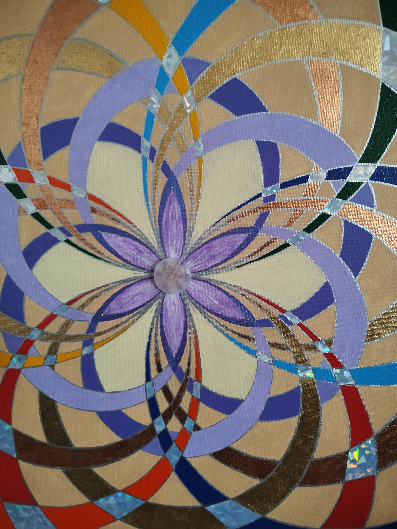 This is a close-up, overhead, studio photograph of a piece of abstract art. The artwork is painted on canvas with a textured surface, appearing to be a mandala-like design. The central element is a stylized purple flower with a clear, round bead at its center. Radiating outward from the center are concentric, curved bands of color in shades of lavender, purple, tan, light yellow, blue, orange, red, brown, and metallic gold and copper. The design is further enhanced with thin silver outlines and small, iridescent, triangular accents scattered throughout the pattern. The background of the canvas is a light tan color. There are no people, objects, or environmental details present in the image, as it focuses solely on the artwork.