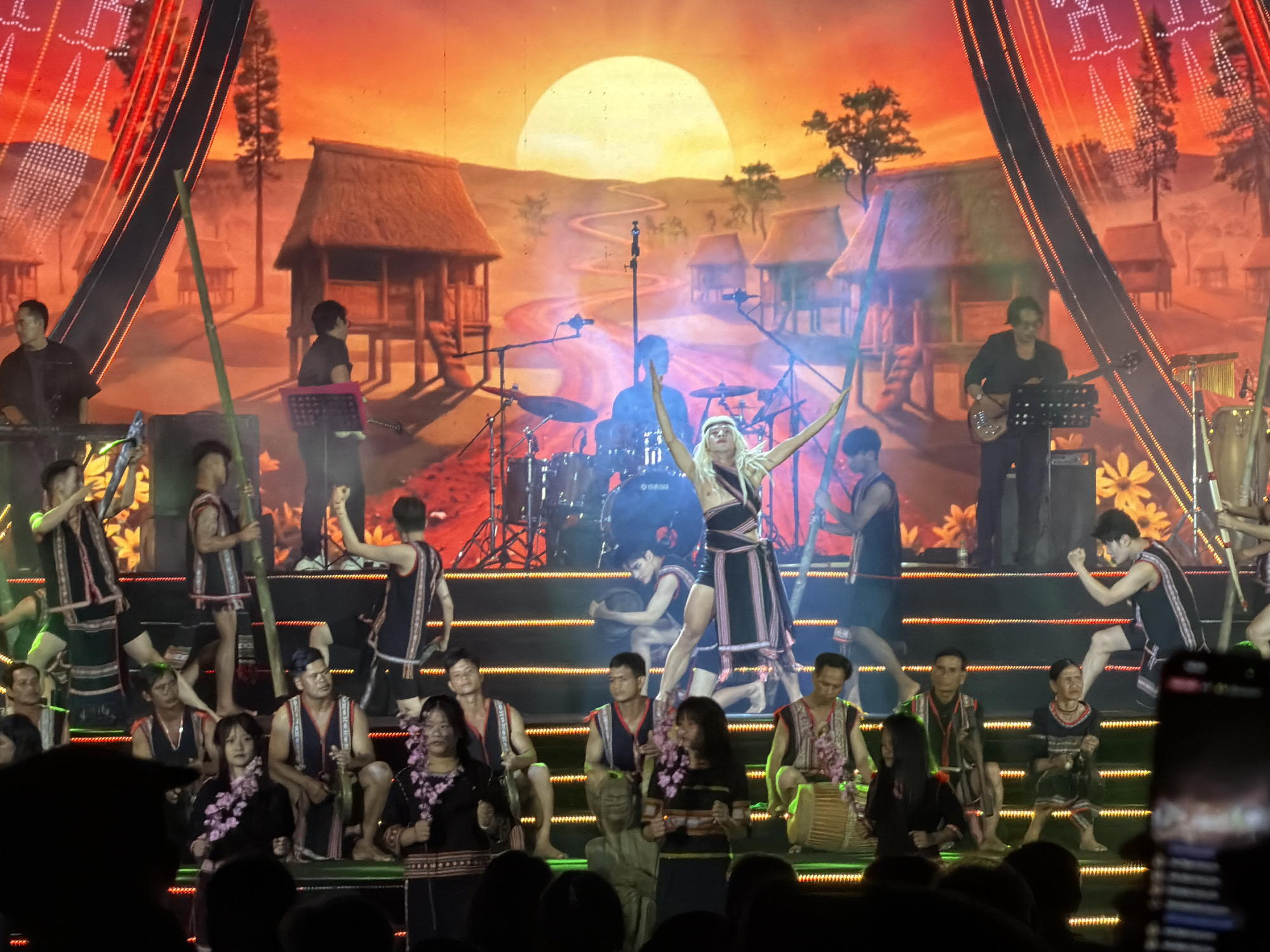 This media file captures a vibrant cultural performance on a multi-level stage, likely in Măng Đen, Vietnam. The backdrop features a painted scene of a traditional village with thatched-roof huts on stilts, winding paths, trees, and a large, glowing yellow sun or moon in the sky.
Numerous performers, dressed in traditional attire with intricate geometric patterns in red, white, and black, are engaged in the show. A central female performer with long blonde hair stands out, arms raised in a dynamic pose, suggesting she is dancing or leading the performance. Musicians are visible, including a drummer with "YAMAHA" faintly written on the drum, and two guitarists/bassists on either side of the stage. Other performers hold long bamboo poles, some dancing, while others are seated on the lower stage levels, playing hand drums.
The stage is brightly lit, indicating an evening performance, and the atmosphere appears energetic and celebratory. Silhouetted audience members are visible in the foreground, with one holding up a phone to record the event. The upper corners of the backdrop feature decorative patterns, complementing the cultural theme.