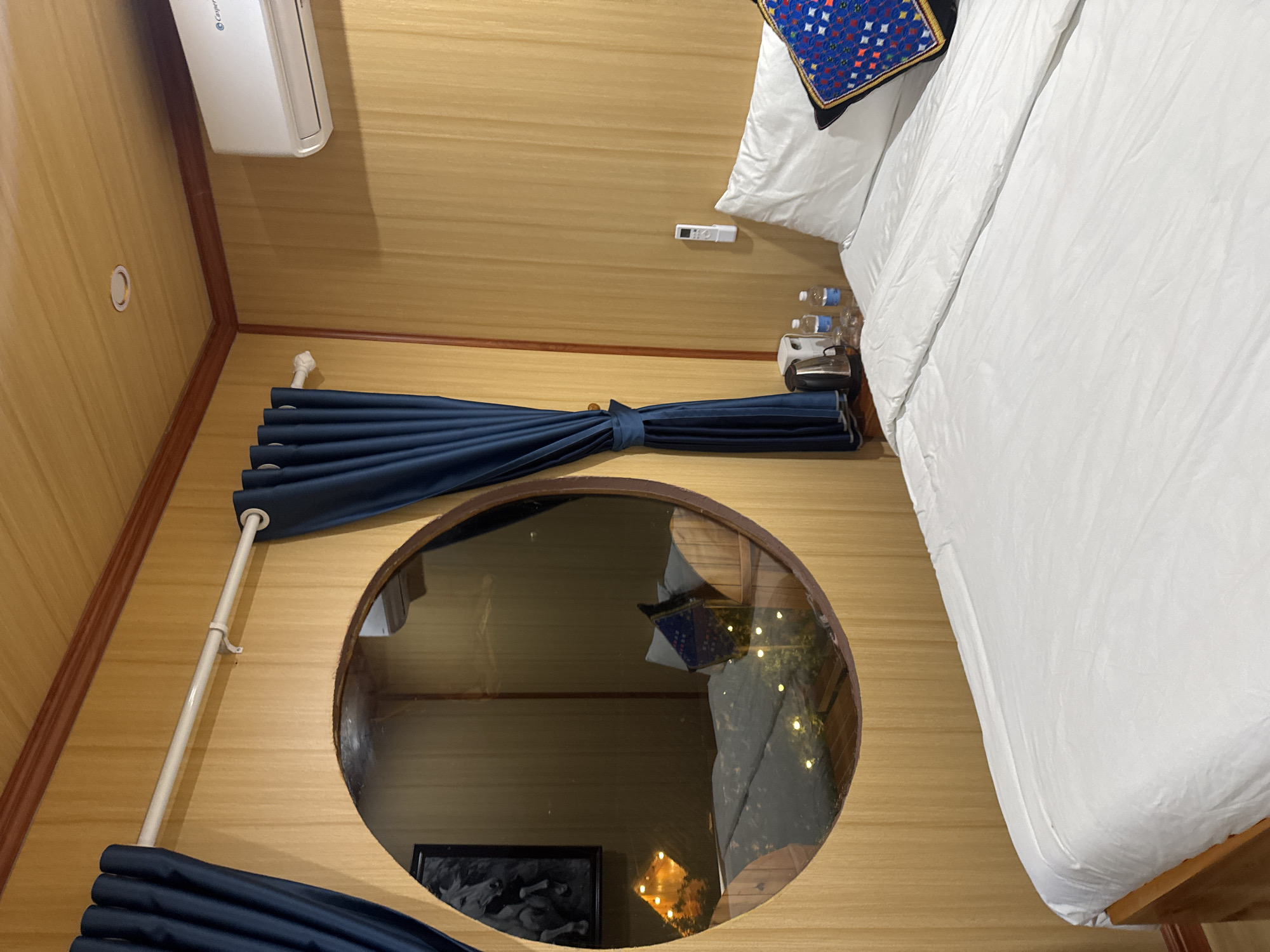 This interior view shows a room in a location in Bắc Hà, Vietnam. The room is paneled with horizontally-aligned wooden boards, and appears to be rotated, with the visible elements including a bed with white linens, and a blue-patterned pillow. An air conditioning unit is affixed to the upper left of the wall. A blue, gathered curtain runs horizontally across the image. A large, circular porthole-style window shows a reflection of the bed and a painting or photograph framed on the wall below the window. A white curtain rod supports the curtain. A remote control and bottled water sits on a nightstand next to the bed.