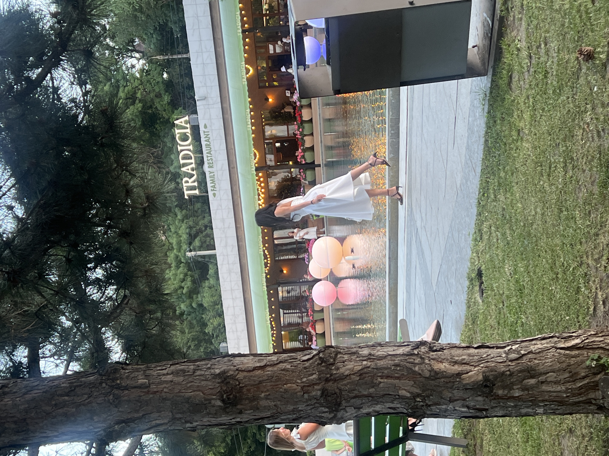 In Odesa, Ukraine, a scene unfolds in front of the "Tradicia Family Restaurant." A large tree trunk dominates the foreground on the left, with its bark textured and aged. Beyond the tree, a woman in a white dress with ruffled shoulder straps stands near a green bench, looking to her left. To her right, a woman in a white dress with thin straps and high heels is captured mid-stride, her body turned to the side as she walks towards the right of the frame. She appears to be walking on a paved area next to a body of water. In the water, large, translucent pink and orange spheres are floating. The restaurant itself is visible in the background, with its green awning and illuminated windows. A sign above the awning reads "TRADICIA" in large, illuminated letters, with "FAMILY RESTAURANT" below it. The environment appears to be an outdoor dining or public space, possibly a promenade or park area adjacent to the restaurant, with lush green grass and trees surrounding the scene.