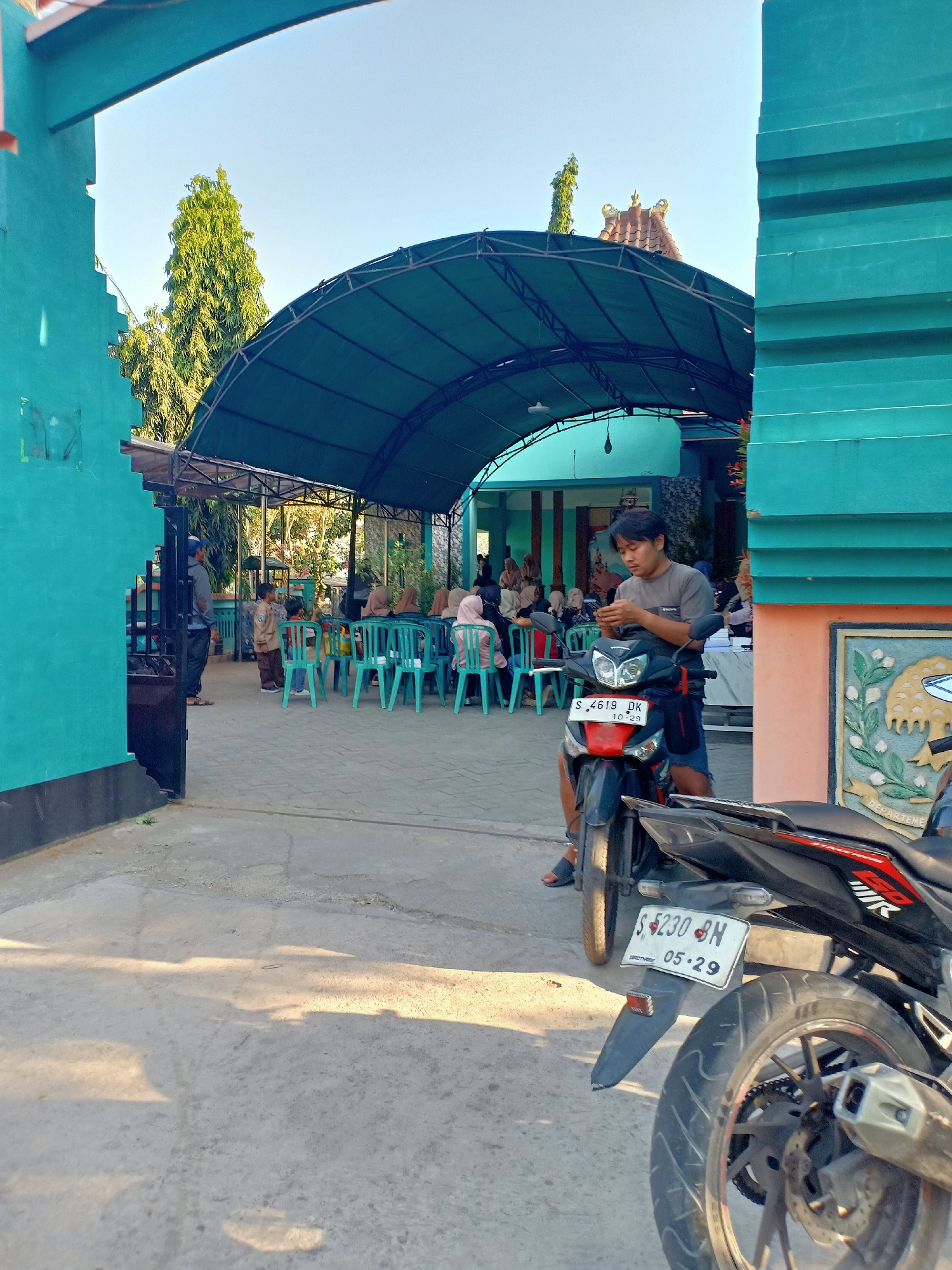 A young man is seated on a motorcycle, looking at his phone. The motorcycle is parked on a paved area, with another motorcycle in the foreground, partially visible. In the background, several people are seated on chairs under a large, green awning. The scene appears to be outdoors, possibly at an event or gathering. The architecture suggests a tropical or warm climate.