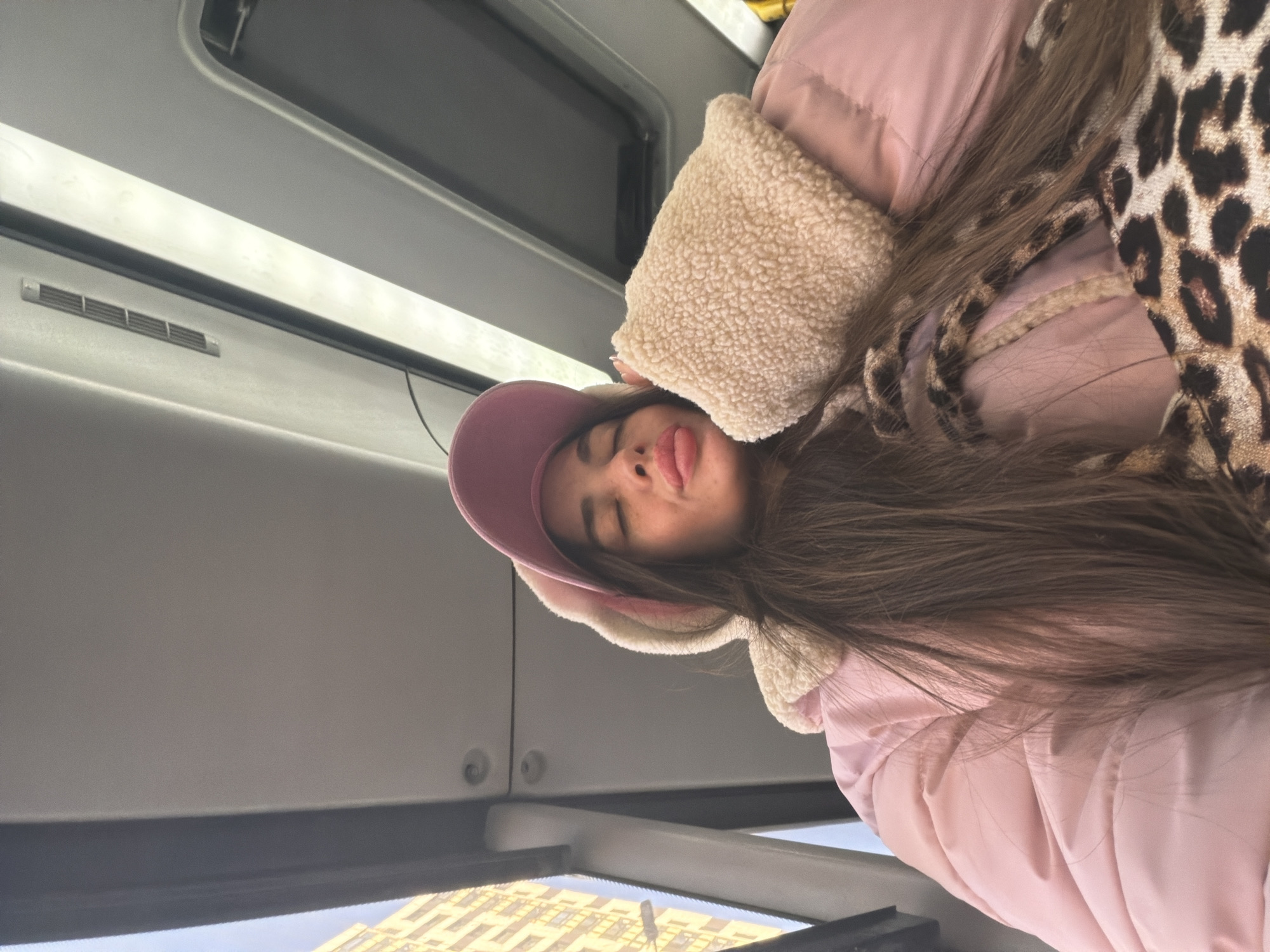 A young woman in a pink jacket and a pink baseball cap with fur trim is seated inside what appears to be a public transportation vehicle, possibly a bus or train. She has long brown hair and is sticking her tongue out slightly. A leopard-print scarf is draped around her neck and shoulders. The interior of the vehicle features a window with a view of a building with yellow walls and multiple windows, which is an indicator of being in the city of Saint Petersburg, Russia. The overhead compartment and side panels of the vehicle are visible, indicating an indoor setting.