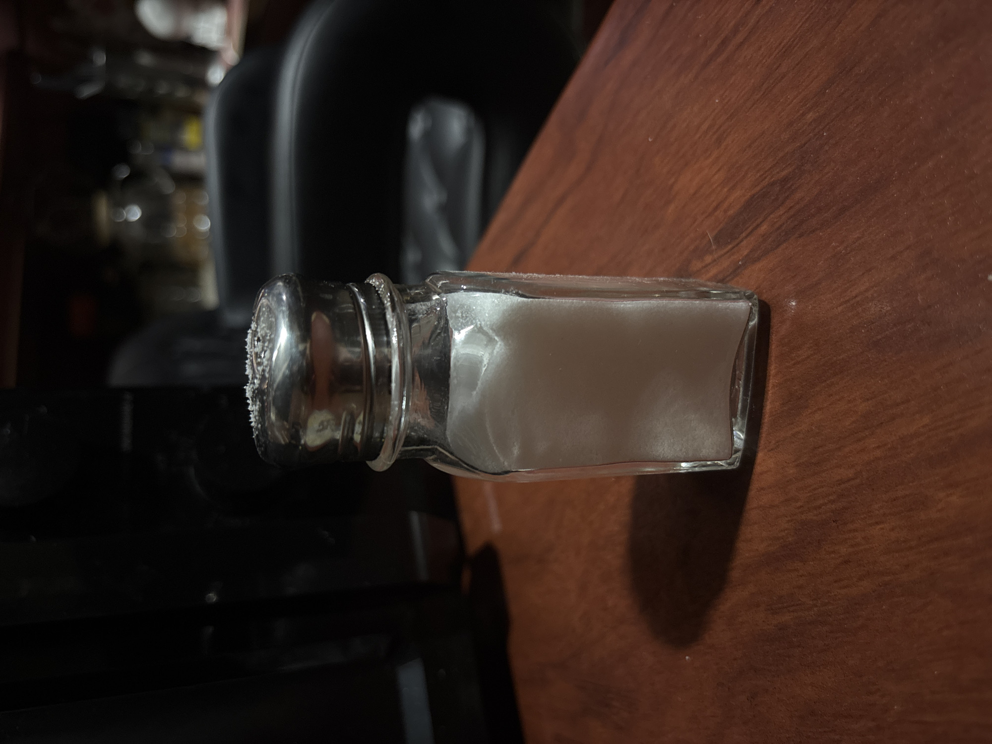 A close-up, horizontal, eye-level shot shows a glass salt shaker filled with white salt. The salt shaker is positioned on a brown wooden surface. The salt shaker has a silver metal cap, which appears slightly corroded or dirty, with a few specks of salt visible on its surface. The glass body of the shaker is clear and rectangular. The salt inside is granular and takes up most of the volume of the shaker.
The background is dark and out of focus. To the left of the salt shaker, a dark, curved shape, possibly the back of a chair or furniture, is visible. Further in the background, indistinct shapes and blurry lights suggest an indoor setting, perhaps a room with other objects or furniture. The lighting appears to be artificial and somewhat dim, casting a shadow of the salt shaker onto the wooden surface. The overall mood of the image is neutral, with no strong emotional cues. There is no visible text in the image. The time of day and weather conditions are not discernible from the provided image.