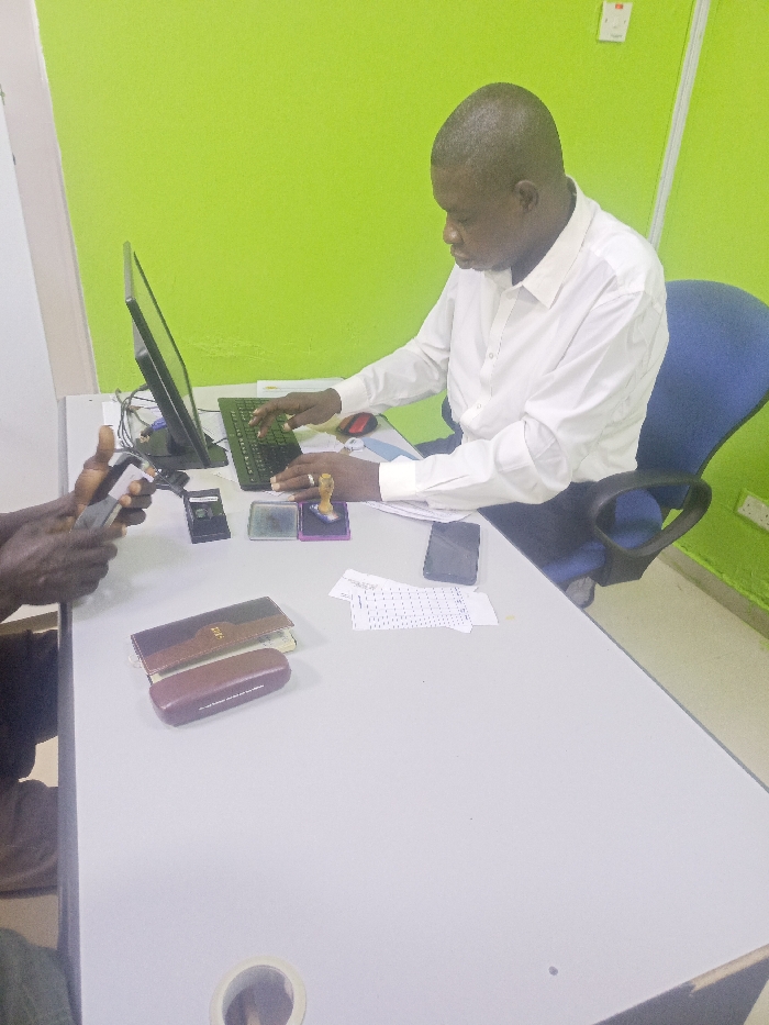 The image depicts an indoor office environment in Akwanga, Nigeria. A light-colored desk with a lime green wall as a backdrop occupies the foreground and midground. On the right, an adult male with short hair, wearing a white long-sleeved shirt and dark trousers, is seated in a blue office chair. He is actively typing on a black computer keyboard, facing right in profile, with a black computer monitor positioned in front of him. A ring is visible on his left ring finger. To the right of the keyboard, a red computer mouse rests on a blue mouse pad.
On the left side of the desk, only the hands and forearms of a second individual are visible, holding and interacting with a black smartphone. Between the two individuals, various objects are arranged on the desk: a brown leather-bound book, a brown glasses case, a black smartphone lying flat, several small pieces of white paper with black markings, a small black square electronic device (possibly a card reader), two ink pads (one transparent, one with a purple tint), and a wooden-handled rubber stamp resting on the purple-tinted pad. A white electrical outlet is visible on the green wall in the upper right. A desk grommet is partially visible in the lower-left foreground.