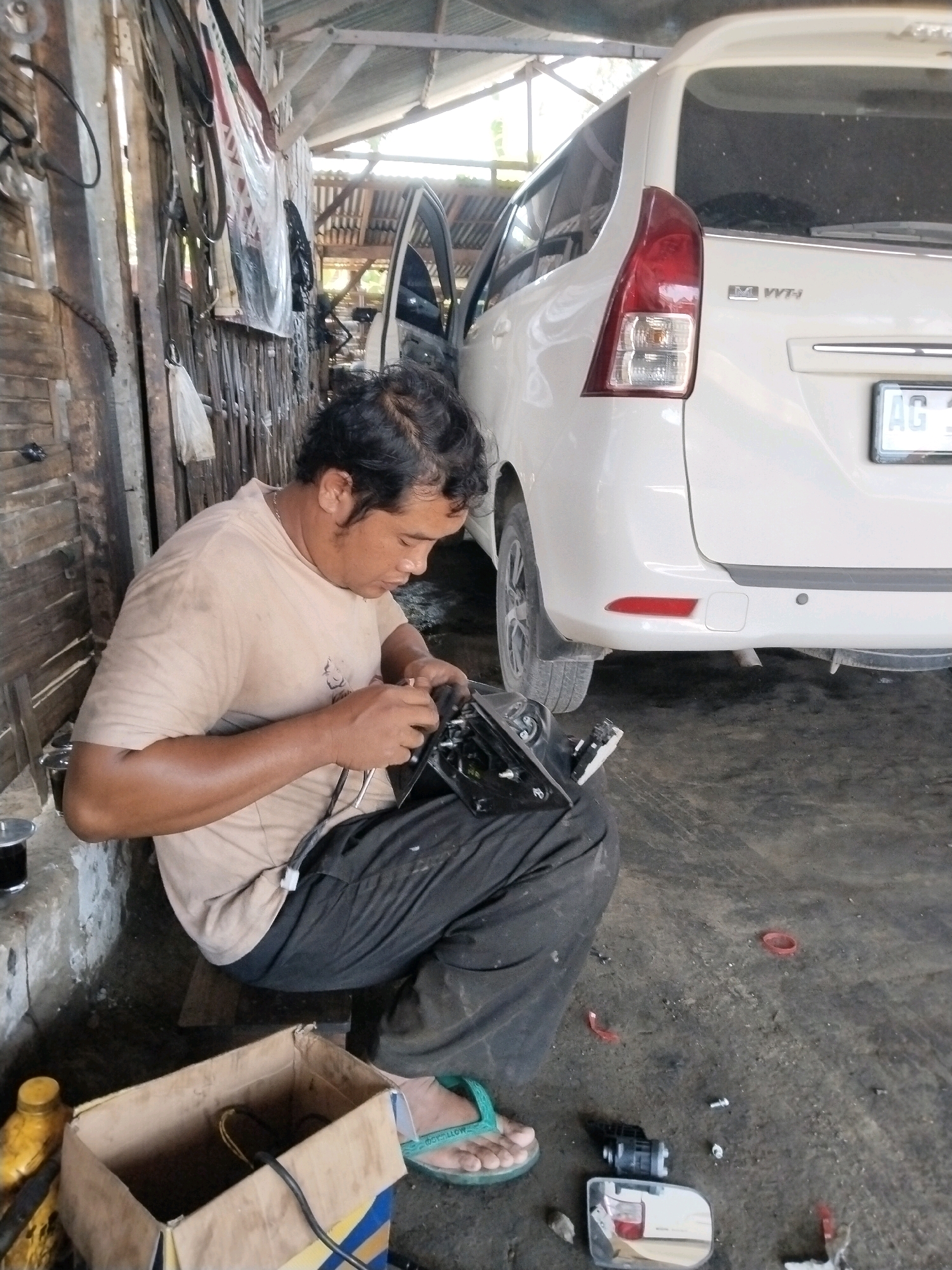 A medium shot shows a man working on a car part inside a garage in Bandung, Indonesia. The man, wearing a light brown t-shirt and black pants, is seated on the floor and focused on a black car mirror assembly he holds in his hands. He appears to be using a tool, possibly pliers, to adjust or connect wires to the assembly. Next to him on the floor is a discarded cardboard box. In the background, a white car is parked, with its rear end visible. The garage environment is rustic, with walls made of wood or bamboo, and a corrugated metal roof. The floor is concrete and appears somewhat dirty, with scattered debris and a small yellow bottle in the foreground. The overall impression is that of an active auto repair workshop.