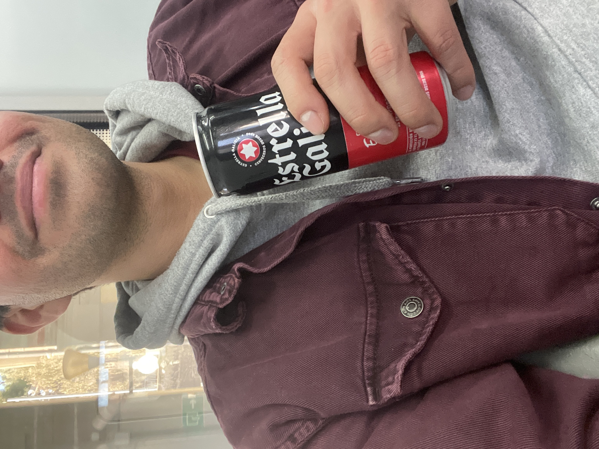 A person is holding a can of Estrella Galicia beer. The can is black and red, with the Estrella Galicia logo prominently displayed. The person is wearing a gray hoodie and a maroon jacket. The scene is inside, possibly near a window, as indicated by reflections. The location is not Caldes d'Estrac, Spain.