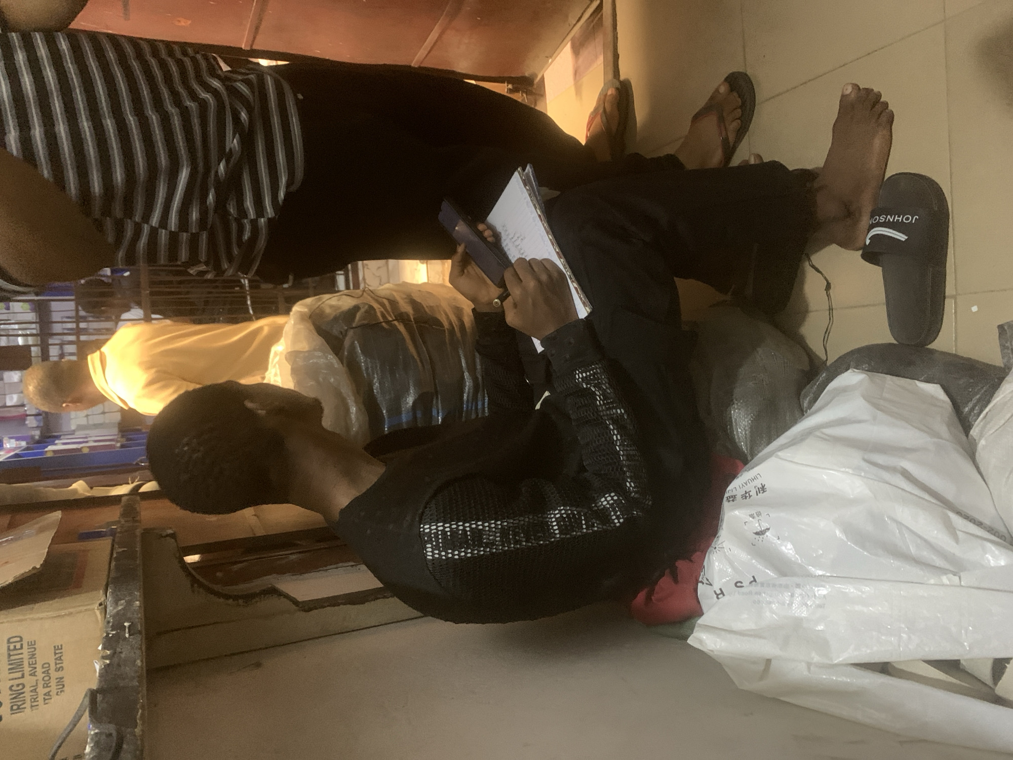A person in a black shirt with a mesh pattern is sitting on the floor, writing in a notebook. They are surrounded by several large, bundled bags, some white and one black, which appear to be containing goods. Another person, wearing a yellow shirt, is partially visible in the background, along with some other people and objects in the distance. In the foreground, a box with the words "IRING LIMITED," "TRIAL, AVENUE," "ITA ROAD," and "GUN STATE" is visible, suggesting a business or commercial setting, located in Fegge, Nigeria. The scene suggests the individual is taking inventory or managing paperwork related to the packed goods around them. One foot is bare, with a black sandal labeled "JOHNSON" nearby.