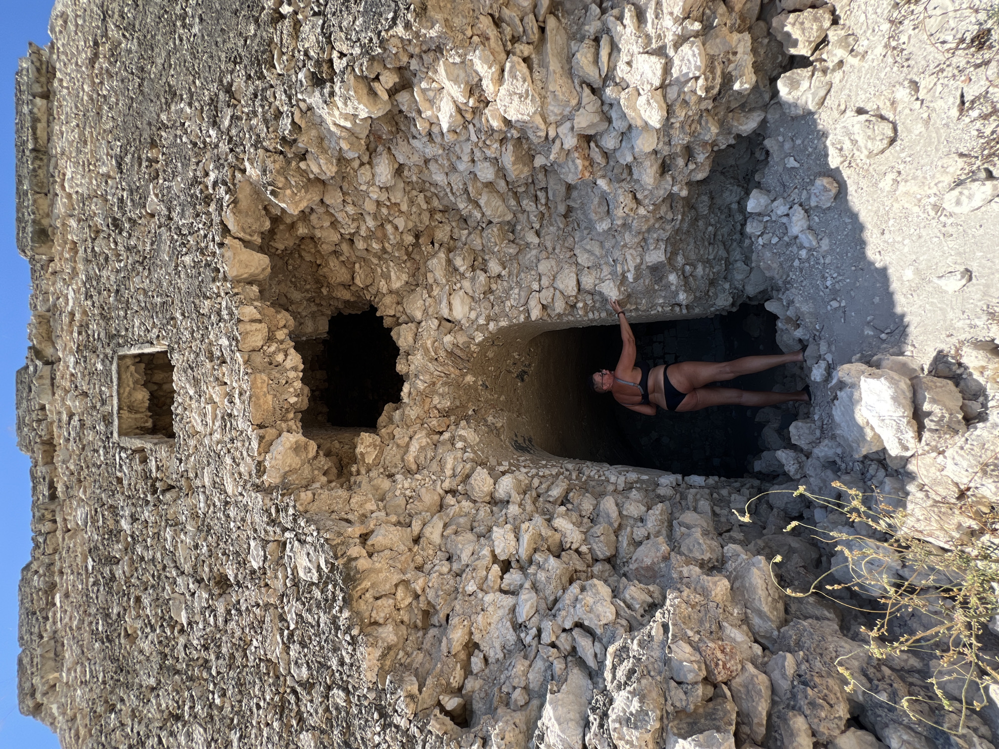 The image shows a woman in a black bikini standing at the entrance of a cave-like opening in a stone structure. The structure appears to be ancient and is made of rough-hewn stones. The opening is arched and leads into a dark interior. The woman is facing away from the camera, looking into the opening and touching the stone wall with her left hand. The ground in front of the opening is covered with rocks and sand. The sky is blue and clear. The location is identified as the Trabucco di San Lorenzo in Peschici, Italy.