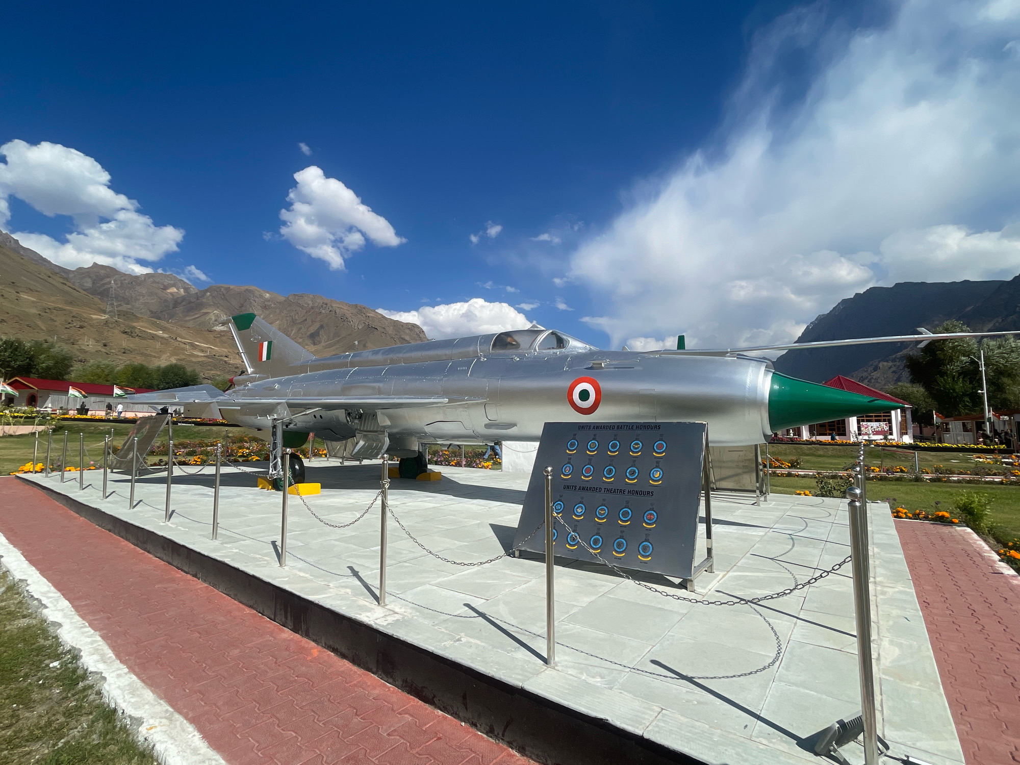 The image displays a static display of a silver fighter jet, likely a Mikoyan-Gurevich MiG-21, placed on a paved platform outdoors. The jet is adorned with the Indian Air Force roundel on its fuselage and the Indian flag on its tail fin. A display board next to the aircraft showcases emblems under the headings "UNITS AWARDED BATTLE HONOURS" and "UNITS AWARDED THEATRE HONOURS." The setting appears to be a memorial or museum in a mountainous region, indicated by the rugged terrain and blue sky with scattered clouds in the background. The foreground features a red brick pathway and green grass. The jet is roped off for public viewing, with metallic posts and chains delineating the exhibition area. No people are prominently visible interacting with the jet. The environment is dry and arid, consistent with a high-altitude region. While the prompt mentions Bhimbat, India, this specific location is not visually confirmed within the provided image; the scene is characteristic of a high-altitude region in India.