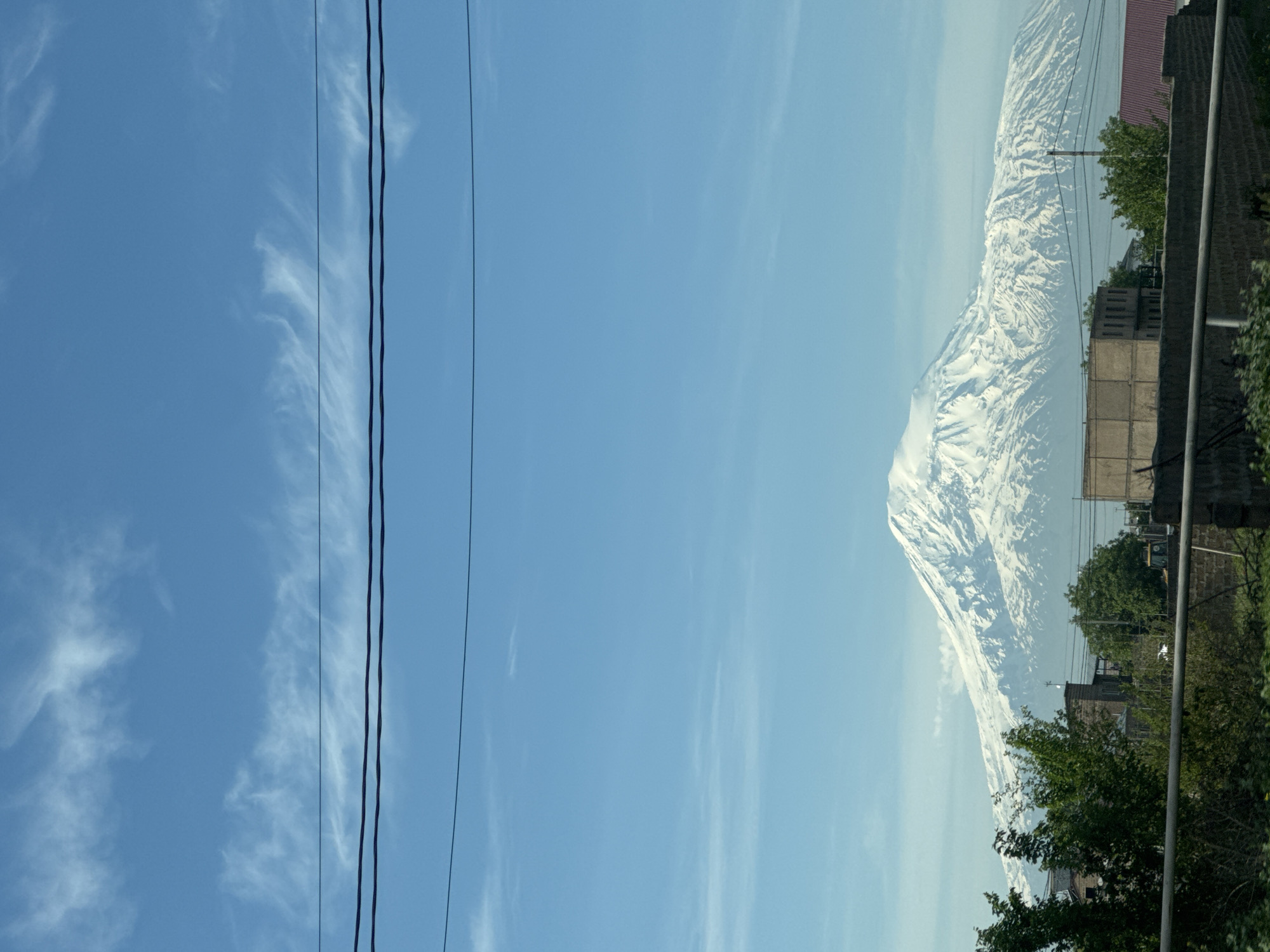 The image shows a view in Musaler, Armenia, featuring Mount Ararat on the right side of the frame. Power lines stretch across the left side against a blue sky with wispy clouds. Buildings and trees are visible at the base of the mountain. The photograph appears to have been taken from a residential area.