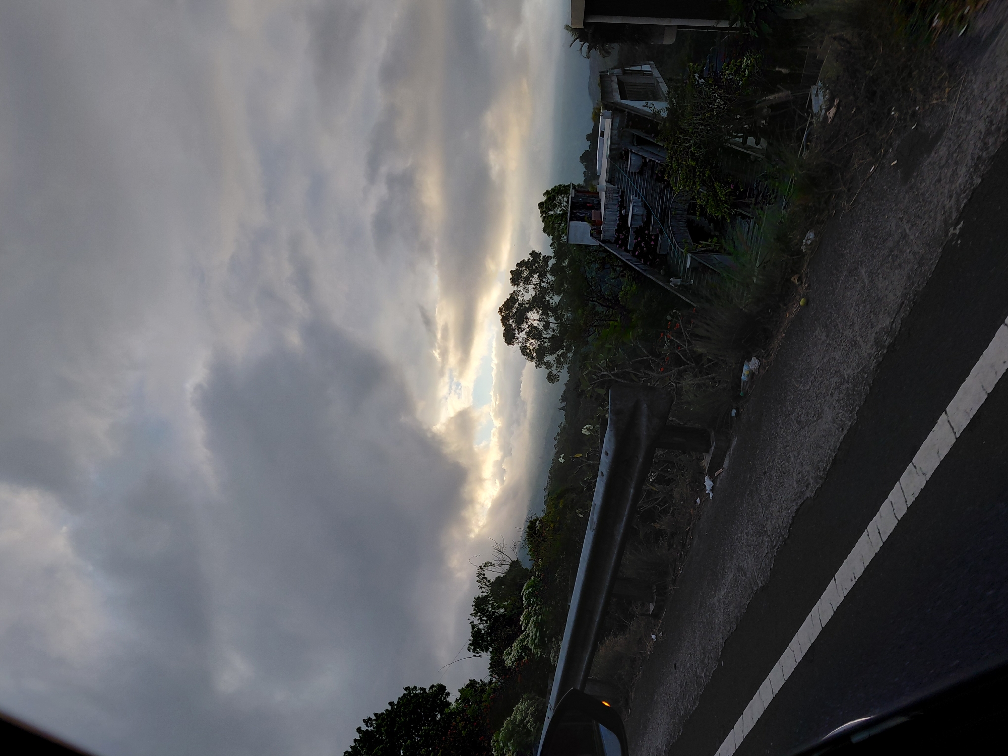 The image depicts a roadside view in Kintamani, Indonesia, characterized by a cloudy sky and a sloped landscape. In the foreground, a paved road with a white line marking is visible, bordered by a metal guardrail. The guardrail appears weathered and is positioned along the edge of a steep embankment.
Beyond the road, the landscape is covered with lush vegetation, including trees and shrubs. In the mid-ground, an unfinished or abandoned building complex is situated on a hillside, featuring multiple levels, staircases, and balconies. Some parts of the structure show signs of disrepair, with debris scattered around. The sky is predominantly overcast, with patches of sunlight breaking through the clouds, creating a dramatic effect over the distant hills. The overall atmosphere suggests a somewhat desolate or neglected location, possibly a former tourist attraction or residential development. There are no people visible in the image.