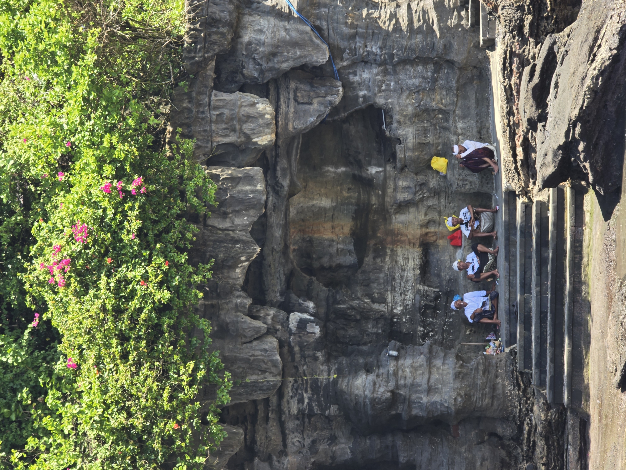 The image captures a scene featuring several individuals amidst a dramatic natural rock formation, complemented by lush greenery. The primary subjects are five men, distinguishable by their white tops, patterned sarongs, and white head coverings, suggesting traditional or ceremonial attire. One man is seated on stone steps in the upper right, while four others are resting or seated near the base of a set of dark stone steps on the lower right side of the frame. Small colorful bags (yellow and red) are visible near some of the men, along with what appear to be small offerings or personal items.
The setting is characterized by a towering, rugged rock cliff face, likely volcanic, exhibiting varied textures and shades of grey and brown. A prominent section of dense, vibrant green foliage, interspersed with bright pink flowers (possibly bougainvillea), covers the left side of the rock face, contrasting sharply with the stone. A blue rope or cable runs vertically down a section of the cliff in the upper middle. The stone steps descend alongside the rock, suggesting access to a lower area.
The men appear to be resting or waiting, perhaps before or after a ritual or gathering, given their attire and composed postures. The scene is brightly lit by natural sunlight, indicating clear weather during the day, likely morning or early afternoon. The overall atmosphere is calm and contemplative. There is no visible text in the image. The combination of traditional attire, the natural rock features, and tropical flora points towards a location in Southeast Asia, possibly an island region known for such landscapes and cultural practices.