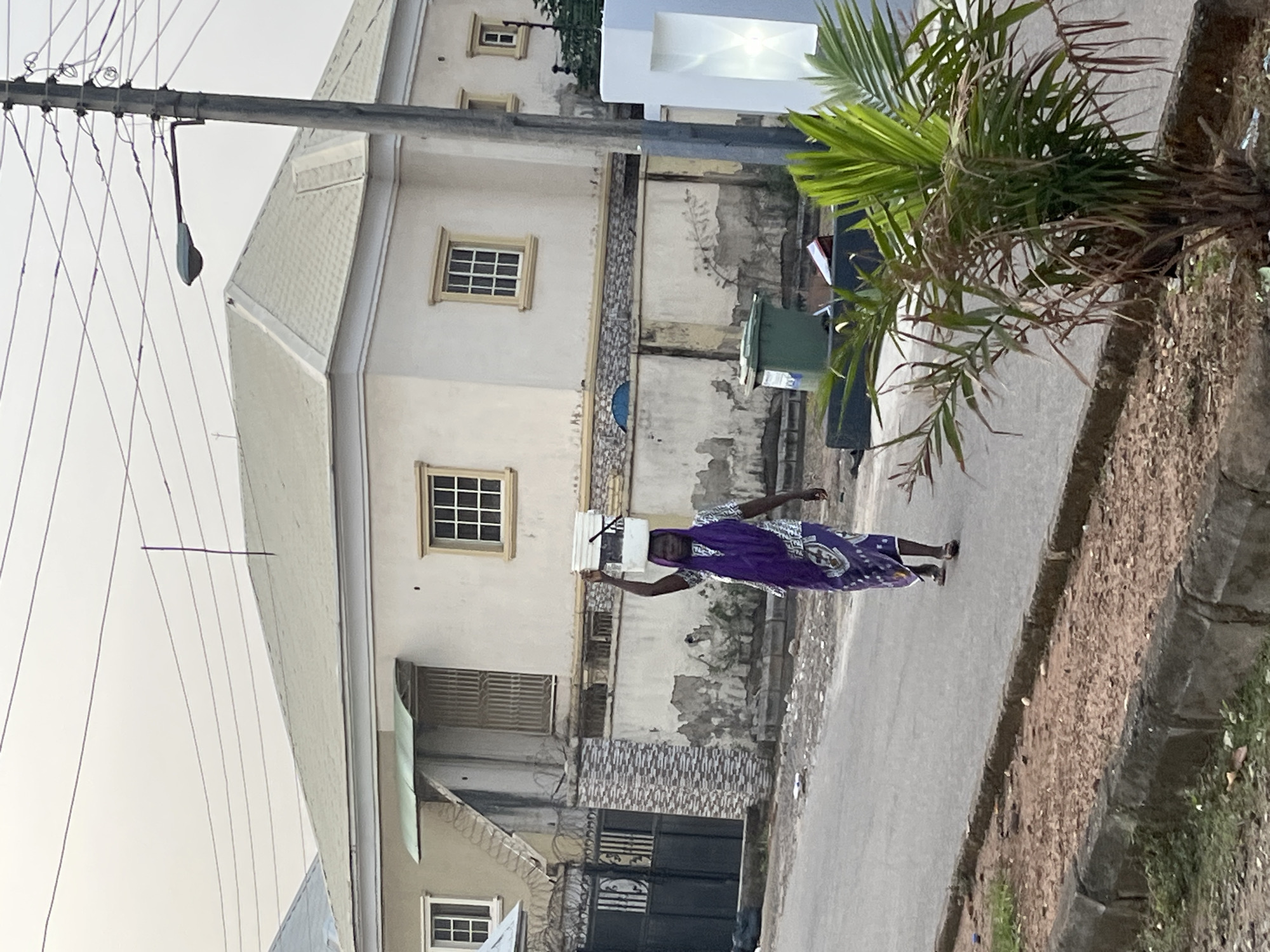 A medium shot shows a woman carrying items on her head while walking down a street in Abuja, Nigeria. The woman is wearing a purple and white patterned garment and sandals. She is walking on a paved road in front of a tall, multi-story building with beige-colored walls and many windows. A green trash receptacle is in front of the building near a small tree. Electrical poles and wires are visible above the building on the left, with vegetation on the right. The overall impression is a typical street scene in a residential or commercial area of Abuja.