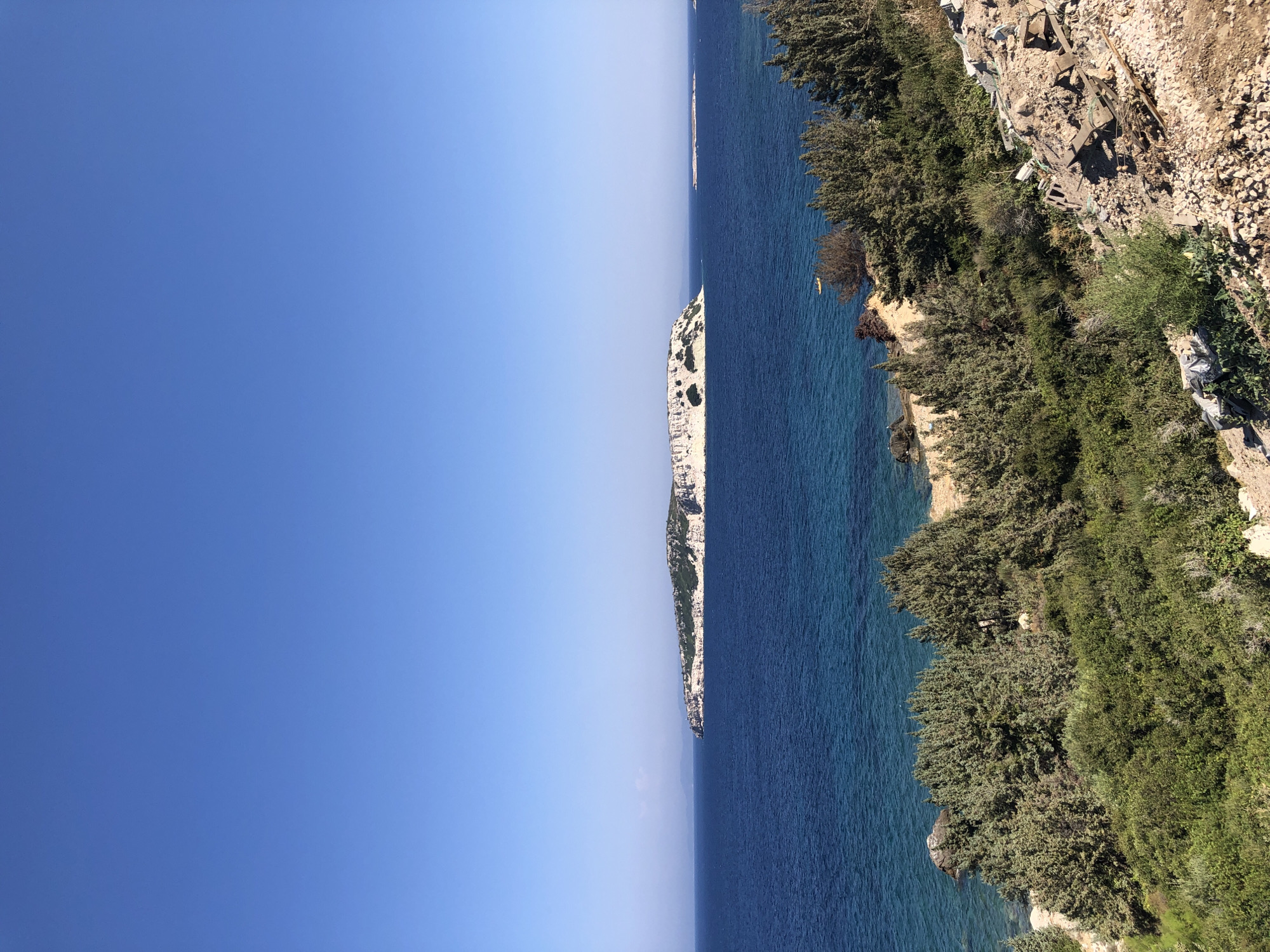 The image shows a panoramic view of the sea and islands near Karaburun, Turkey. The sky is clear and blue, and the sea is a deep blue color with lighter turquoise shades near the shore. In the foreground, there is a rocky, vegetated hillside with several green trees and bushes. The middle ground features the coastline with more vegetation and a rocky beach. In the background, there are two islands. The larger island in the center is flat-topped with light-colored cliffs and green vegetation. A smaller island is visible to the right of the larger one. The water around the islands appears calm. There is a small yellow object, possibly a boat or kayak, in the water between the foreground vegetation and the large island. The overall scene is natural and serene.