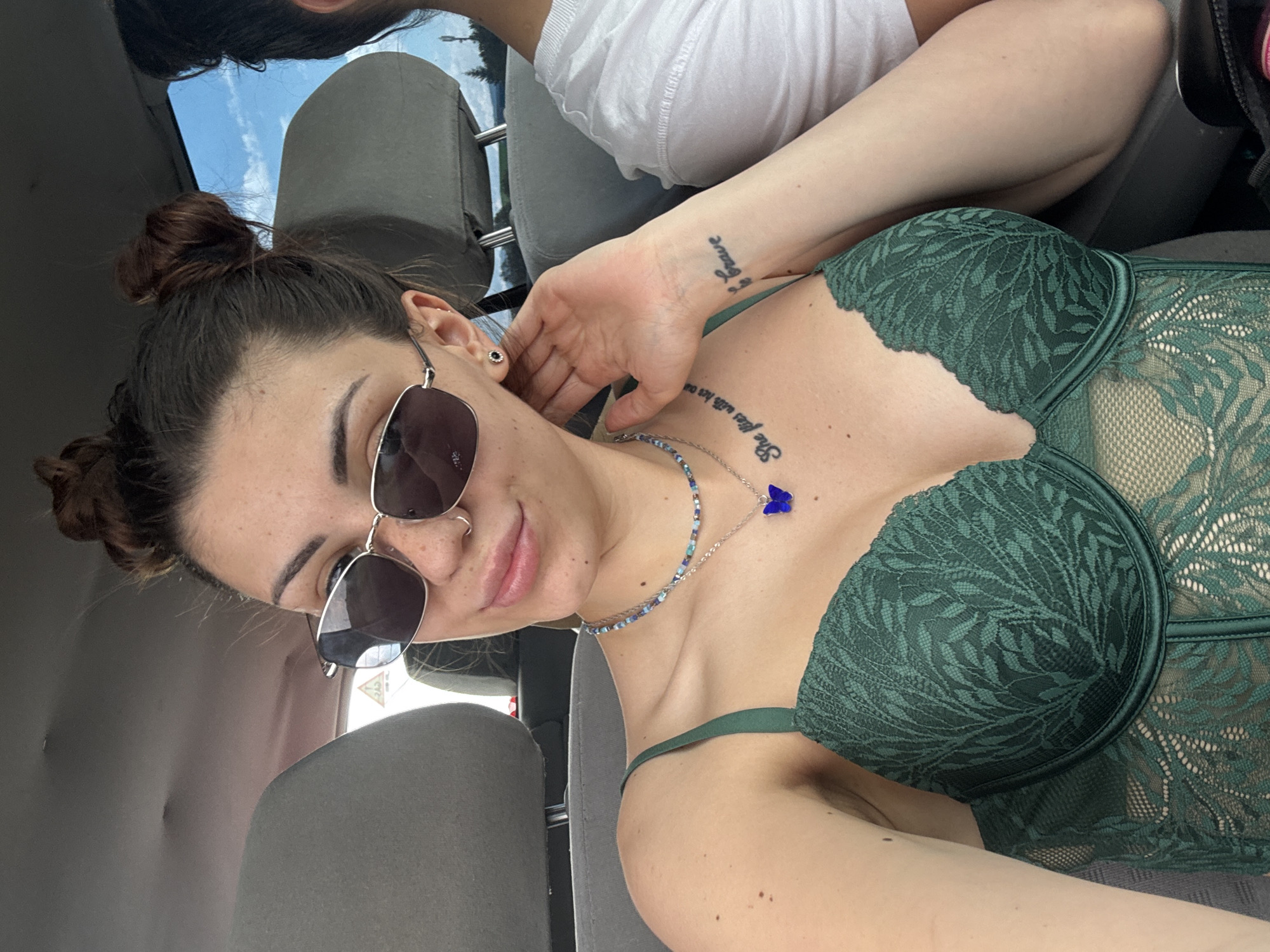 An overhead shot captured inside a car in Gorna Malina, Bulgaria, shows a young woman in the foreground, angled to her left, looking towards the camera with a slight smile. She has dark hair styled in two small buns and is wearing sunglasses. Her left hand is raised towards her face, and a small black earring is visible. She wears a dark green lace top or bodysuit with intricate patterns. Around her neck are two necklaces: one beaded with blue and green tones, and a silver chain with a blue butterfly pendant. A tattoo on her upper chest/collarbone area reads "She flies with her ou" (incomplete). On her right wrist, another tattoo appears to say "Brave". Freckles are visible on her nose and cheeks, along with a nose ring. Partially visible in the top left corner is another person, possibly a passenger, wearing a white t-shirt, with their arm extended towards the foreground woman. The car's interior, including grey seats and headrests, is visible. Through the car window, a bright blue sky with some clouds is seen, indicating daytime.