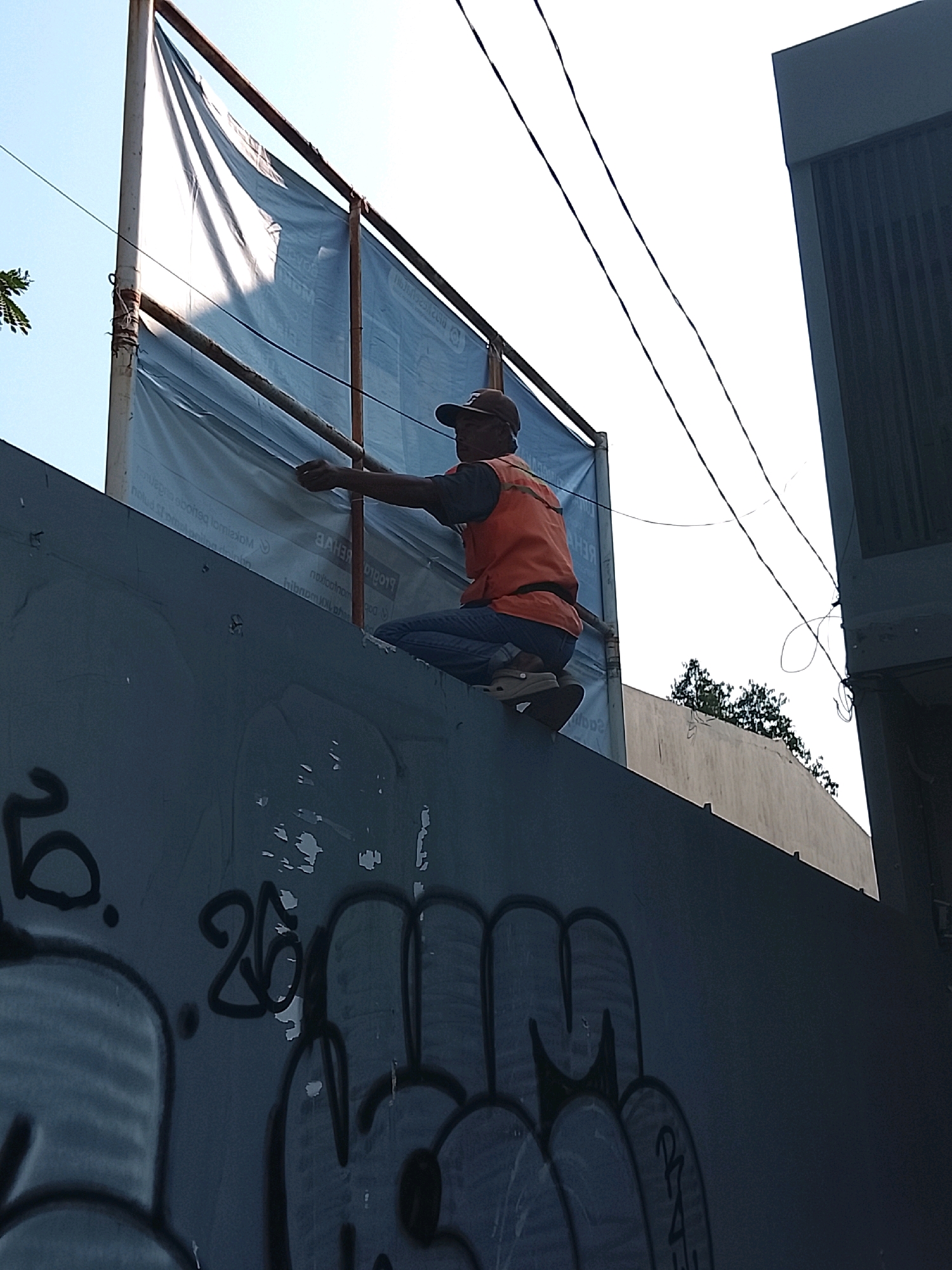 A person wearing an orange vest and a cap is crouching on top of a dark gray wall, appearing to be working on a large blue banner attached to a metal frame. The banner is somewhat obscured by shadows and sunlight, and it has visible text printed on it. Power lines are visible in the sky above the banner and the person. To the right of the banner is a dark-colored building with vertical slats. The wall the person is on is covered in graffiti, with visible tags and drawings in black and gray. The overall environment suggests an outdoor urban setting, possibly a construction or maintenance site. The location is identified as Slawi, Indonesia.