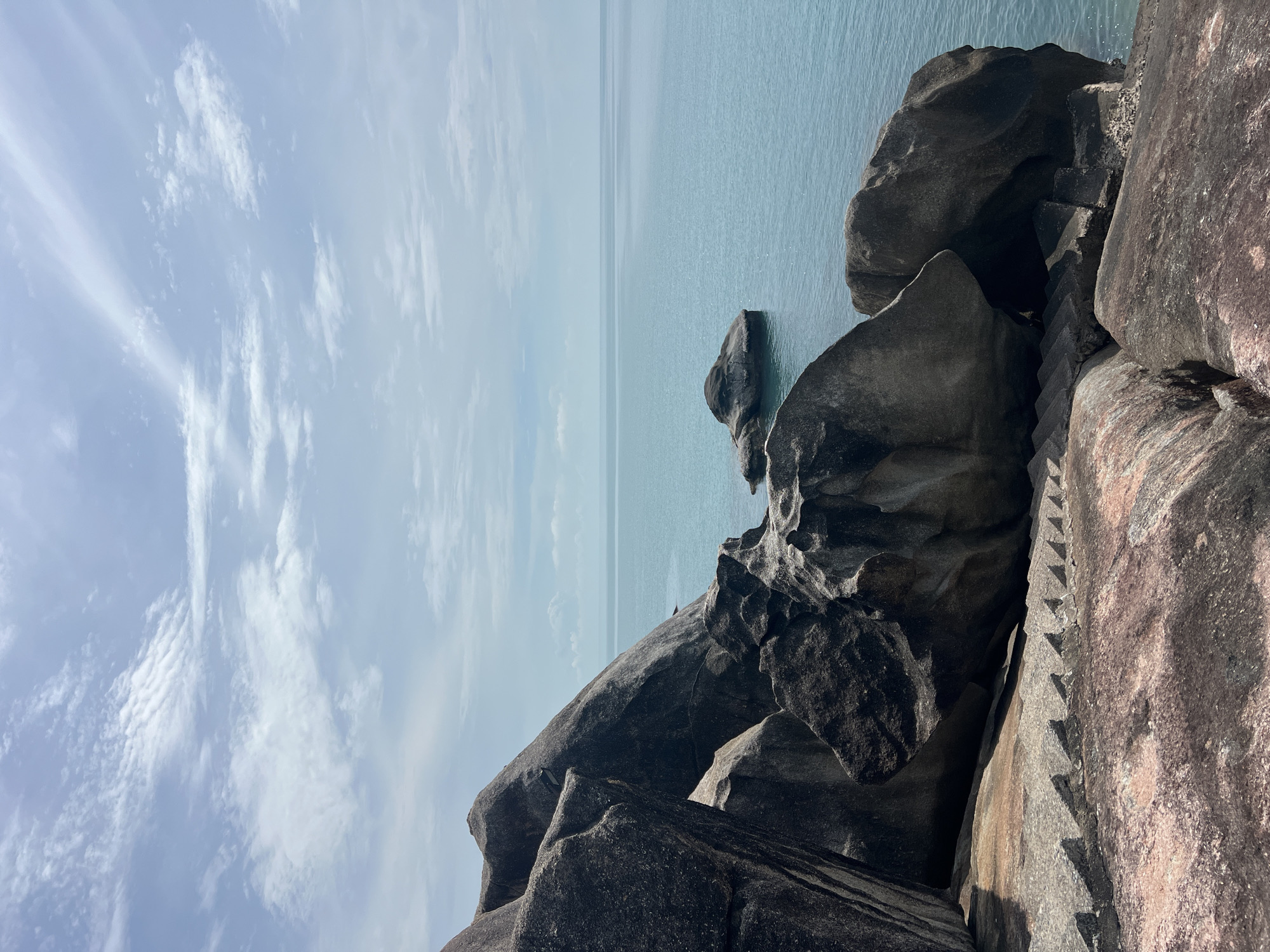 An eye-level, vertical, medium shot captures a coastal landscape in Baan Chaweng Noi, Thailand. In the foreground, weathered, large granite boulders, with pinkish and gray hues, dominate the scene. A set of stone steps is carved into the rock face, descending towards the water. These steps are partially obscured by the immense rocks. Beyond the rocks, the turquoise ocean stretches to the horizon under a pale blue sky dotted with wispy white clouds. A few smaller rocks emerge from the water in the mid-ground, adding to the rugged beauty of the location. The overall mood is serene and natural, with no people present.