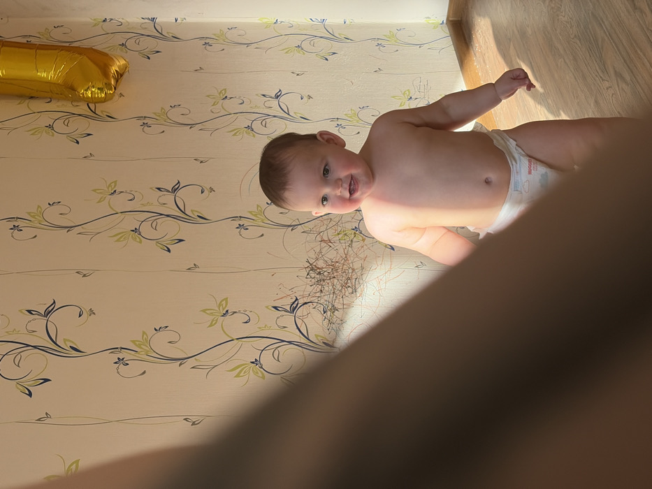 The image depicts an indoor scene within a domestic residence in the city of Chaikovsky, Russia. A fair-skinned infant, appearing approximately one year old, stands in the midground, facing slightly right of the camera. The infant has light brown hair, is bare-chested, and wears a white disposable Huggies diaper with light blue and pink graphics. The infant's mouth is open in a wide smile or laugh, with visible upper and lower teeth/gums. Strong natural sunlight illuminates the infant's right side, the right wall, and the floor, casting a bright patch. The left foreground is occupied by a blurred, dark brown mass, consistent with a partial human arm or shoulder, extending diagonally from the top left to the lower center, obscuring approximately one-third of the frame. The background consists of a wall covered in light-toned wallpaper featuring repeating vertical floral patterns in dark blue and light green. Below one of these patterns, near the center-left, the wallpaper exhibits a dense area of multi-colored scribbles in red, orange, and black. In the upper right corner, a partially visible golden, reflective foil balloon, consistent with the shape of the number "1," is affixed to the wall. The floor is covered with light brown laminate or parquet flooring.
