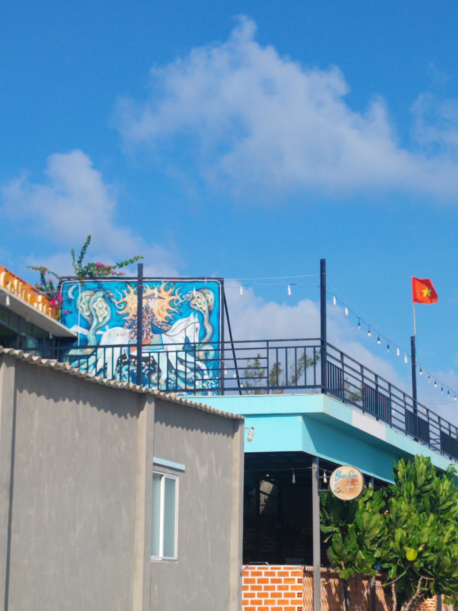 The image depicts a two-story building in Phú Quý Special Administrative Region, Vietnam, under a bright blue sky with scattered clouds. A gray concrete building is in the foreground, partially obscuring the view. Above the gray building, a turquoise-colored structure is visible, featuring a black metal railing and string lights hanging along its perimeter. A Vietnamese flag is attached to one of the posts on the roof. A large, blue-toned mural is mounted on the roof of the building next to the turquoise one, with a design that includes human and equine figures. A 'Blue Sky Cafe' sign hangs near the base of the turquoise building, partially obscured by foliage. The building has orange brick details.