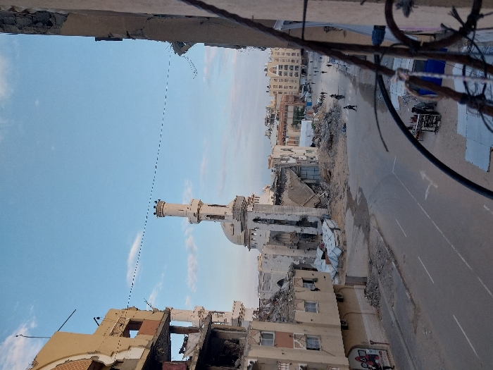 The image captures a wide view of a severely damaged urban landscape, indicative of a conflict zone or the aftermath of a major disaster. The perspective is elevated, looking down over the scene.
In the foreground, the edge of a damaged building or structure from which the photograph was taken is visible. Twisted metal wires and cables hang into the frame from the top right, suggesting further structural damage directly above the viewer's position.
The mid-ground is dominated by a wide, paved road running horizontally across the image. The road has visible white dashed lines and double yellow lines, indicating traffic lanes. Large amounts of dirt, sand, and rubble are scattered along the verges of the road and on its surface in places.
Flanking both sides of the road are numerous buildings, many of which are heavily damaged or completely destroyed. On the left side, structures are reduced to piles of rubble, with some walls partially standing, revealing exposed interiors and broken concrete. On the right side of the road, a prominent mosque with a tall, slender minaret and a dome is visible. The mosque itself shows significant damage, with parts of its facade and structure appearing broken or missing. Adjacent to and behind the mosque are more destroyed buildings and debris.
Further in the background, a density of urban buildings can be seen, some appearing less damaged than those in the immediate vicinity, suggesting a wider city area. The sky above is clear and light blue with a few scattered white clouds, indicating daytime.
A few very small, dark figures are discernible on the distant section of the road; they appear to be people walking or possibly a motorcycle. The overall scene conveys a strong sense of devastation and the profound impact of destructive events on urban infrastructure and life.
No discernible text is clearly visible or legible anywhere in the image.