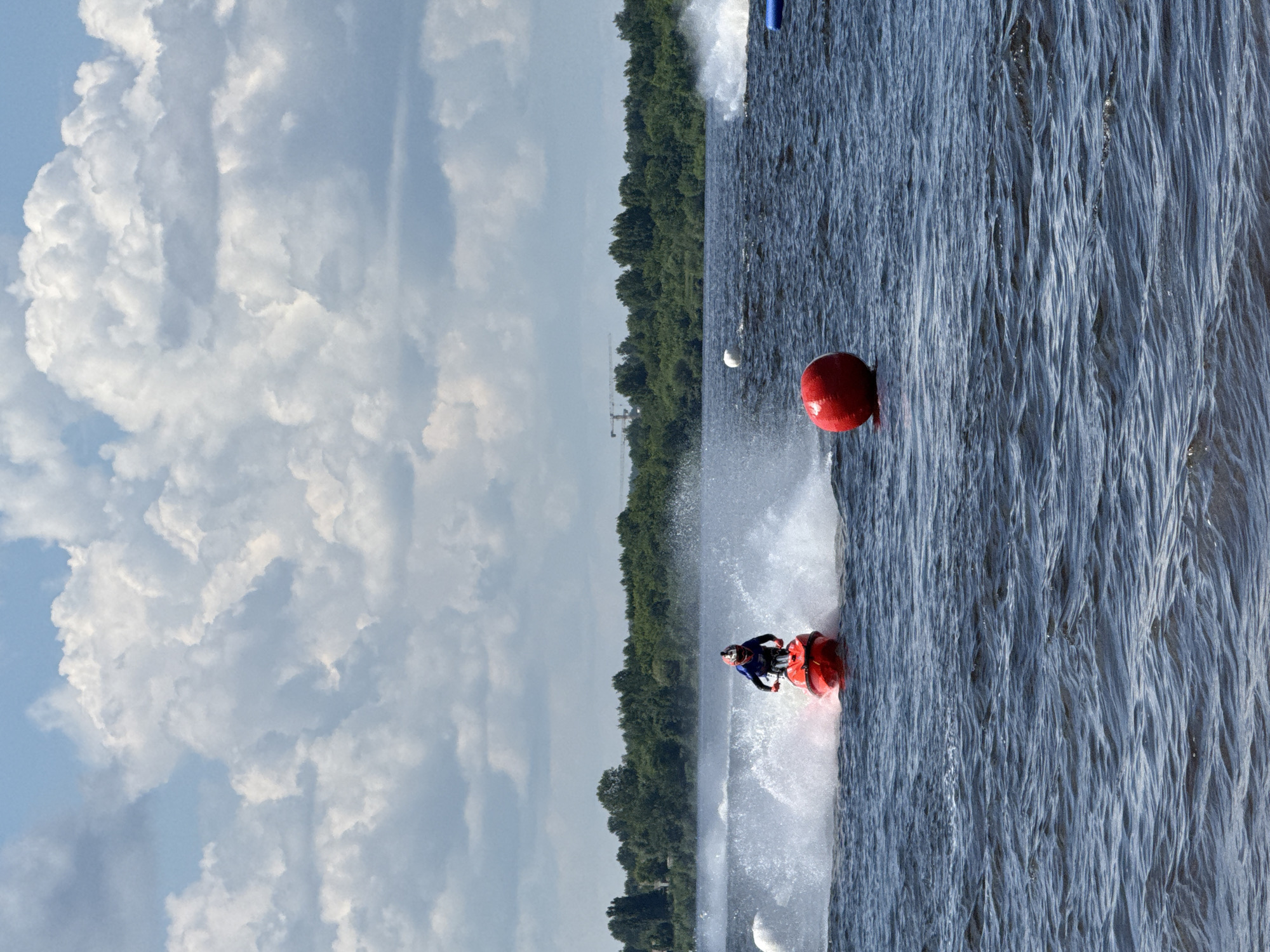 A person rides a red jet ski on a body of water. The rider is wearing a blue shirt and a red helmet. The jet ski is creating a large spray of water. In the background, there is a lush green treeline with some distant construction cranes. The sky is blue with fluffy white clouds. In the foreground, there is a large red buoy floating in the water. The scene appears to be a jet ski race or a recreational activity on the Kama River in Perm, Russia.