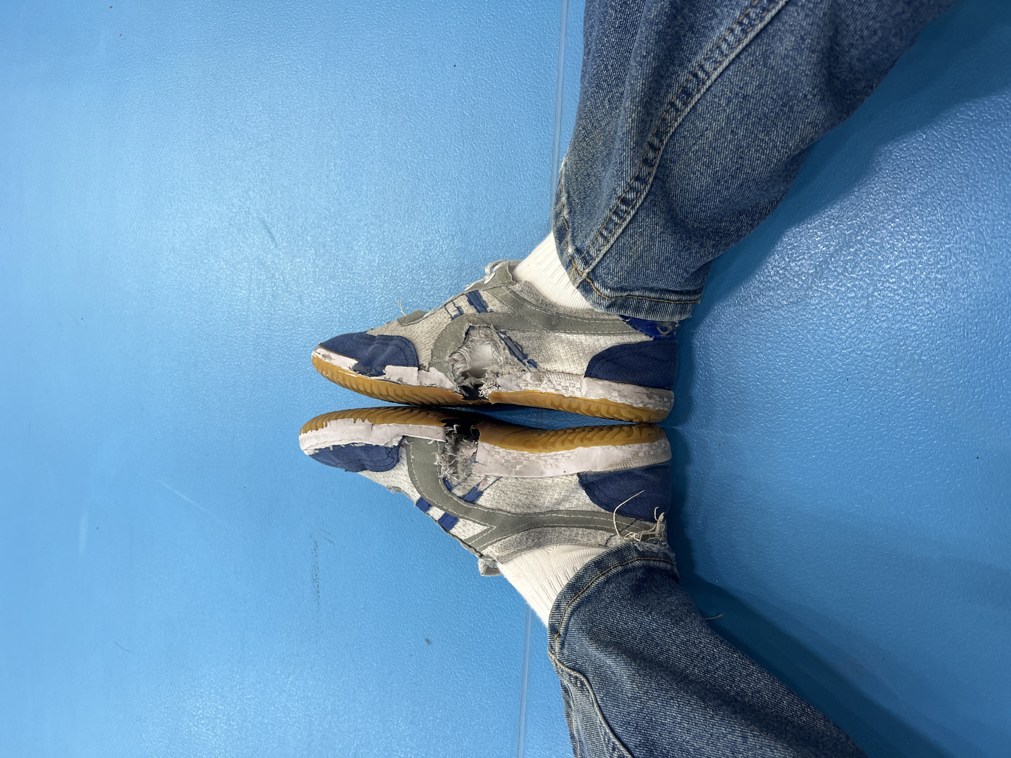 The image shows a top-down view of two feet wearing damaged sneakers on a blue floor. The sneakers are blue, gray, and white with a brown sole, and have large holes on the sides. The feet are wearing white socks and blue jeans. The blue floor appears to be made of a textured material, possibly linoleum or rubber. There is no evidence that this photograph was taken in the city of Berezniki, Russia.