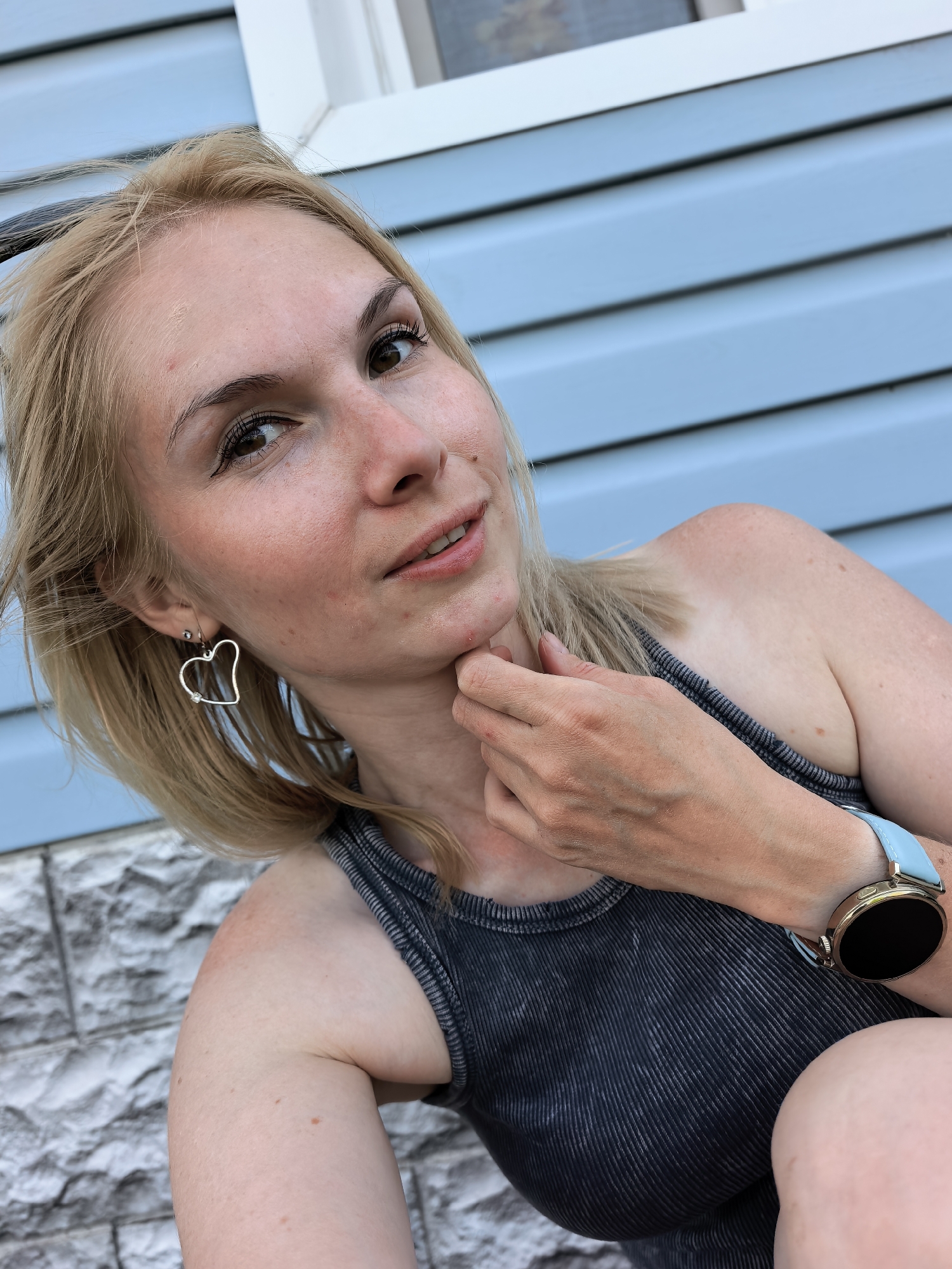 A woman with blonde hair is posing for a selfie. She is wearing a grey tank top and heart-shaped earrings. A smartwatch with a light blue band is visible on her wrist. The background consists of light blue siding and a white windowsill. The location is Sterlitamak, Russia.