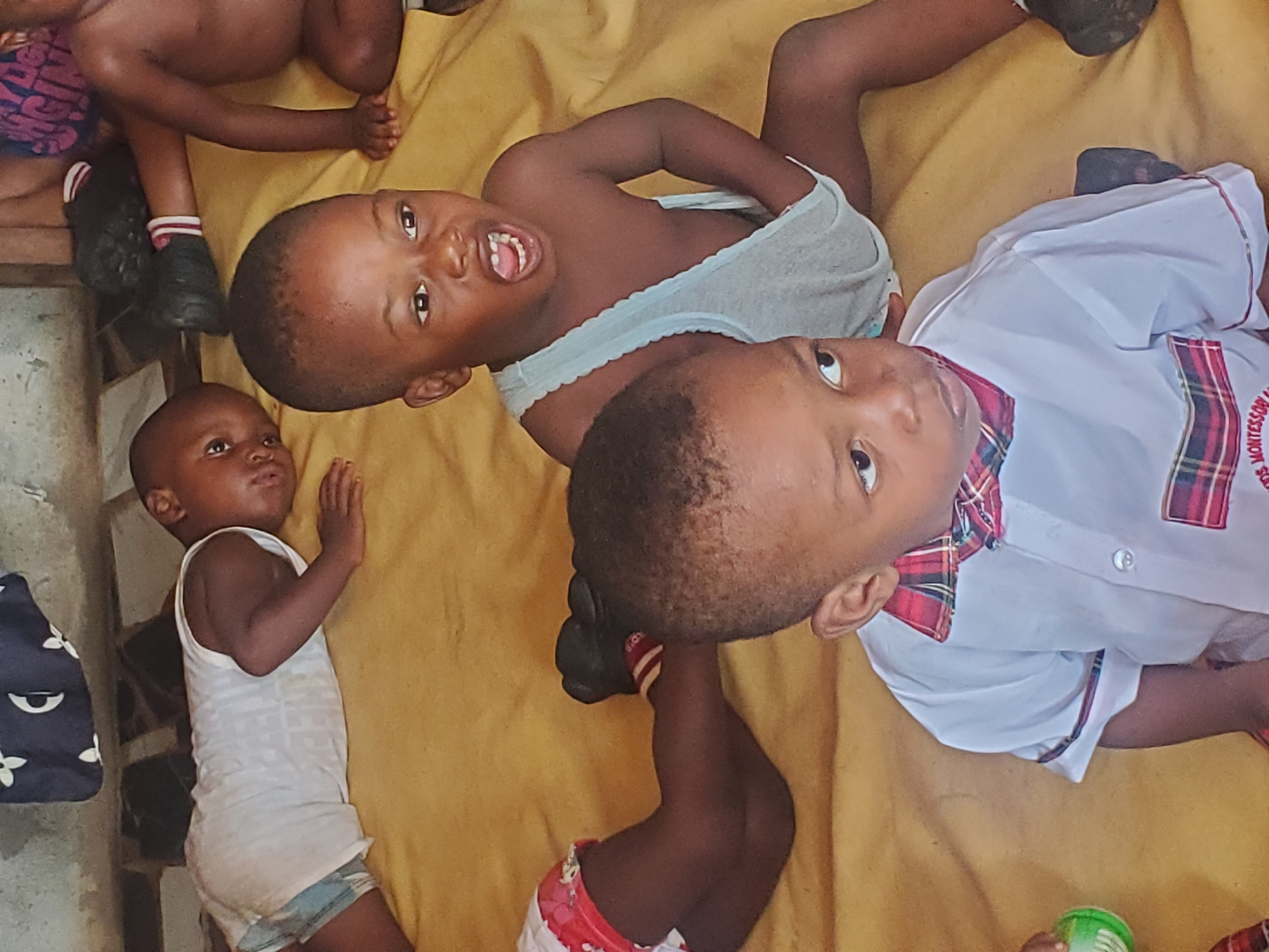 The image depicts a group of young children lying on a yellow cloth or blanket. One child is wearing a white shirt with a plaid collar and a "Montessori" logo on the chest. Another is wearing a gray tank top. One child is wearing a white tank top. The children appear to be playing or resting indoors. The image was taken in Ukwu-Abwa, Nigeria.