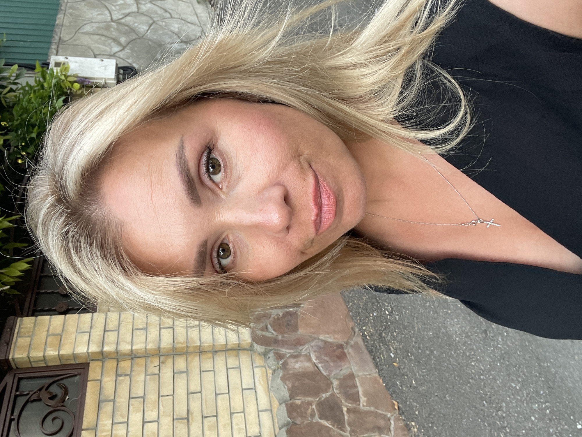 The image is a close-up, rotated, selfie-style photograph of a woman with blonde hair. She is looking at the camera with a slight smile. She has light brown eyes, pink lipstick, and appears to be wearing a black top. A delicate silver necklace with a cross pendant is visible around her neck. The background suggests an outdoor setting with a textured stone or brick wall on the left and what appears to be asphalt or pavement on the right. Green foliage is visible in the upper left portion of the image. The lighting suggests it is daytime. The overall mood is casual and friendly. There is no visible text in the image.