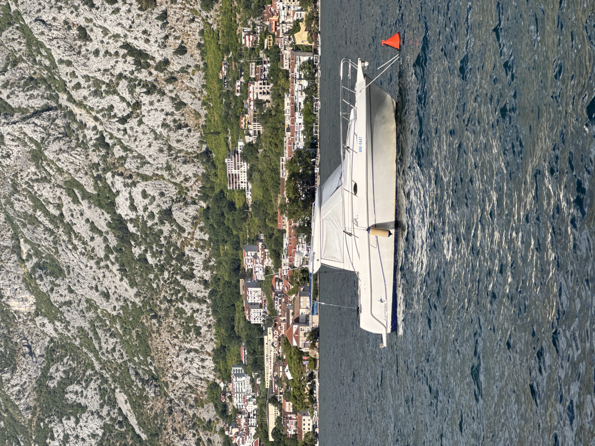 A white motorboat with a covered cabin is anchored in the dark blue waters of the Bay of Kotor, near the coastal town of Prčanj, Montenegro. The boat, which has the registration number MNE-0447 displayed on its hull, is secured by a rope to a bright orange buoy. Behind the boat, the town of Prčanj stretches along the coastline, characterized by red-tiled roofs and a mix of residential buildings and some larger structures. Towering above the town is a steep, rocky mountain face sparsely populated with green vegetation. The scene is bathed in sunlight, indicating a clear day.