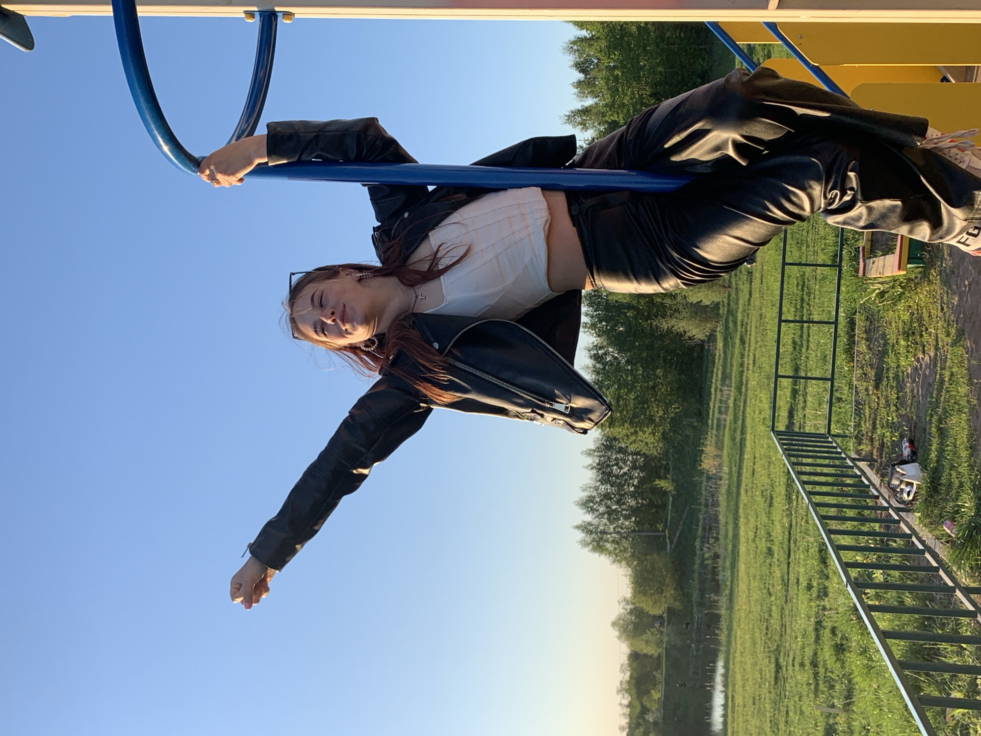 A woman is suspended upside down from a blue playground bar in Фоки, Russia. She is wearing a white top, black leather jacket, and black leather pants. Her right arm is extended downward with her fist clenched. A park with green grass and trees surrounds the playground equipment. A metal ladder leading to the playground structure is visible. The sky is clear and blue.