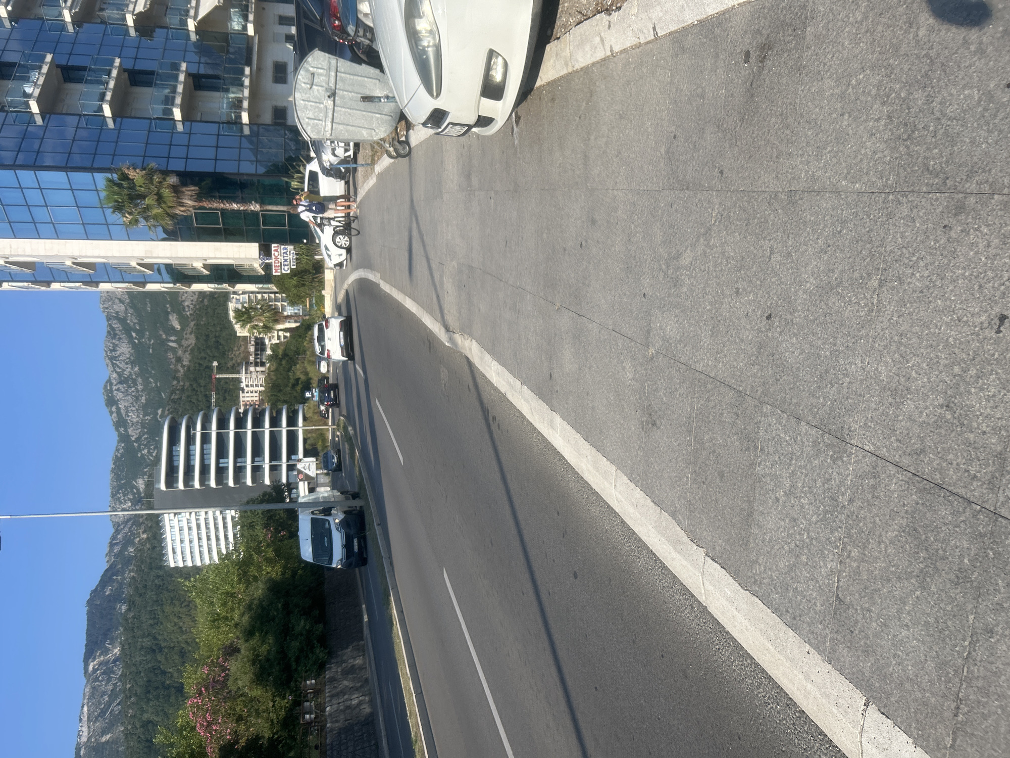 The image shows a street scene in Boreti, Montenegro, on a sunny day. The perspective is from the side of the road, looking towards a modern building with reflective glass windows and balconies, and in the distance, a lush green mountain.
Several vehicles are visible. In the foreground, a white car with its hood open is parked on the side of the road. Further down the road, a white van is parked. There are also glimpses of other cars and a motorcycle. A person is seen near the white car, appearing to be interacting with it or standing next to it. Another person is on a bicycle.
The road has a white line marking its edge. The sidewalk is paved with large, grey tiles. The overall scene suggests a moderately busy urban or coastal area. The presence of mountains in the background and the clear blue sky indicate good weather. The lighting suggests it is daytime, likely late morning or early afternoon due to the strong sunlight and shadows.
A sign is visible on the modern building with text that appears to read "MEDICAL CENTAR". This sign provides a specific detail about the location. The architecture of the building with its curved elements and extensive glass suggests a contemporary design. The foliage, including a palm tree near the building, hints at a warm climate.